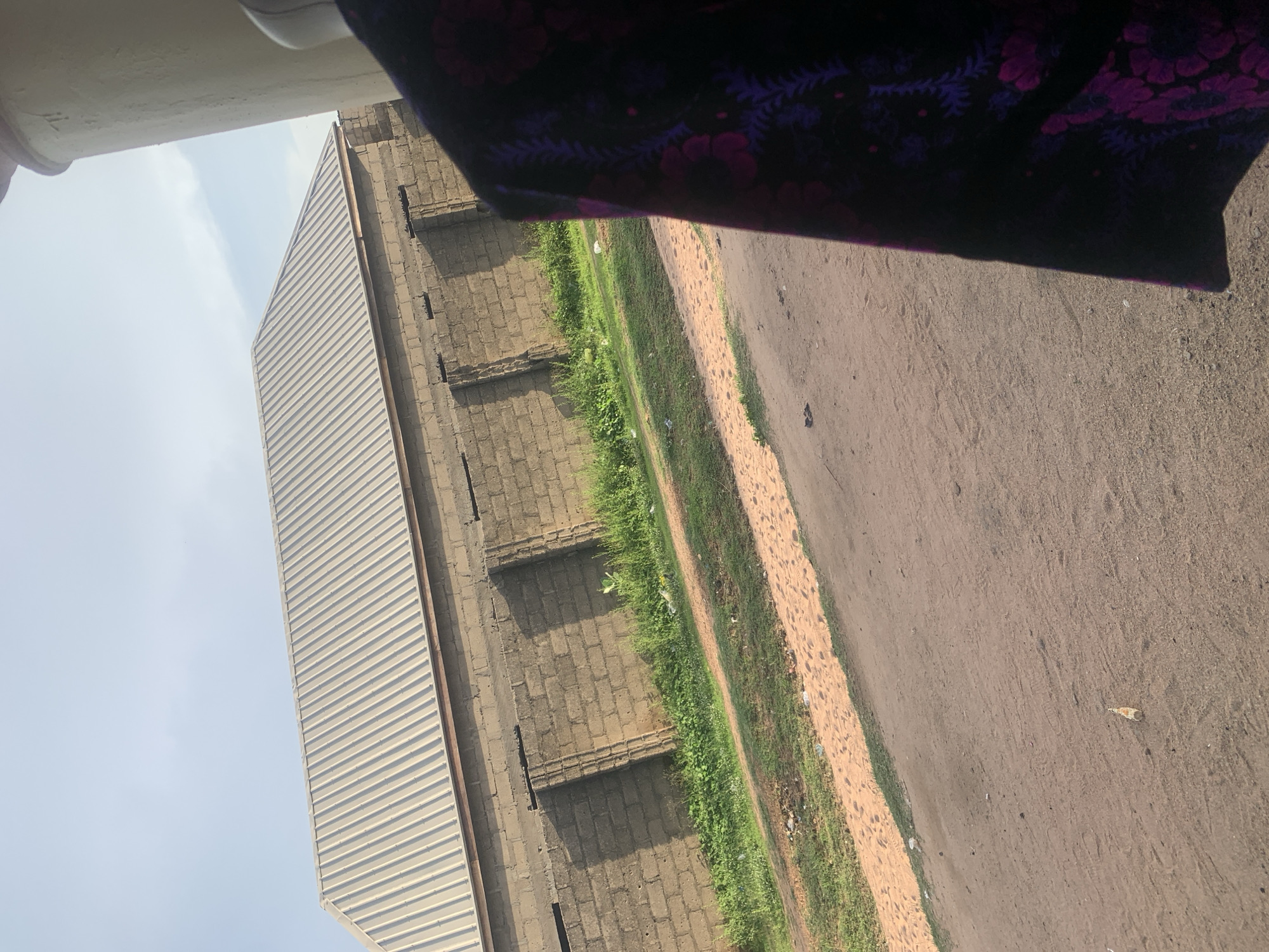 The image depicts an outdoor scene at ground level, capturing a dirt road in the foreground. To the left of the road, there is a strip of green grass interspersed with some scattered white debris, possibly litter. Beyond the grass lies a wall constructed from cinder blocks, with regularly spaced vertical indentations. Surmounting the cinder block wall is a building with a corrugated metal roof that slopes downwards. The sky above is clear with a hint of pale blue. On the far right of the image, a portion of a dark garment with a floral pattern in shades of purple and pink is visible, suggesting the presence of a person or clothing item within the frame. The scene appears to be in an unpaved area, likely a rural or semi-urban setting.