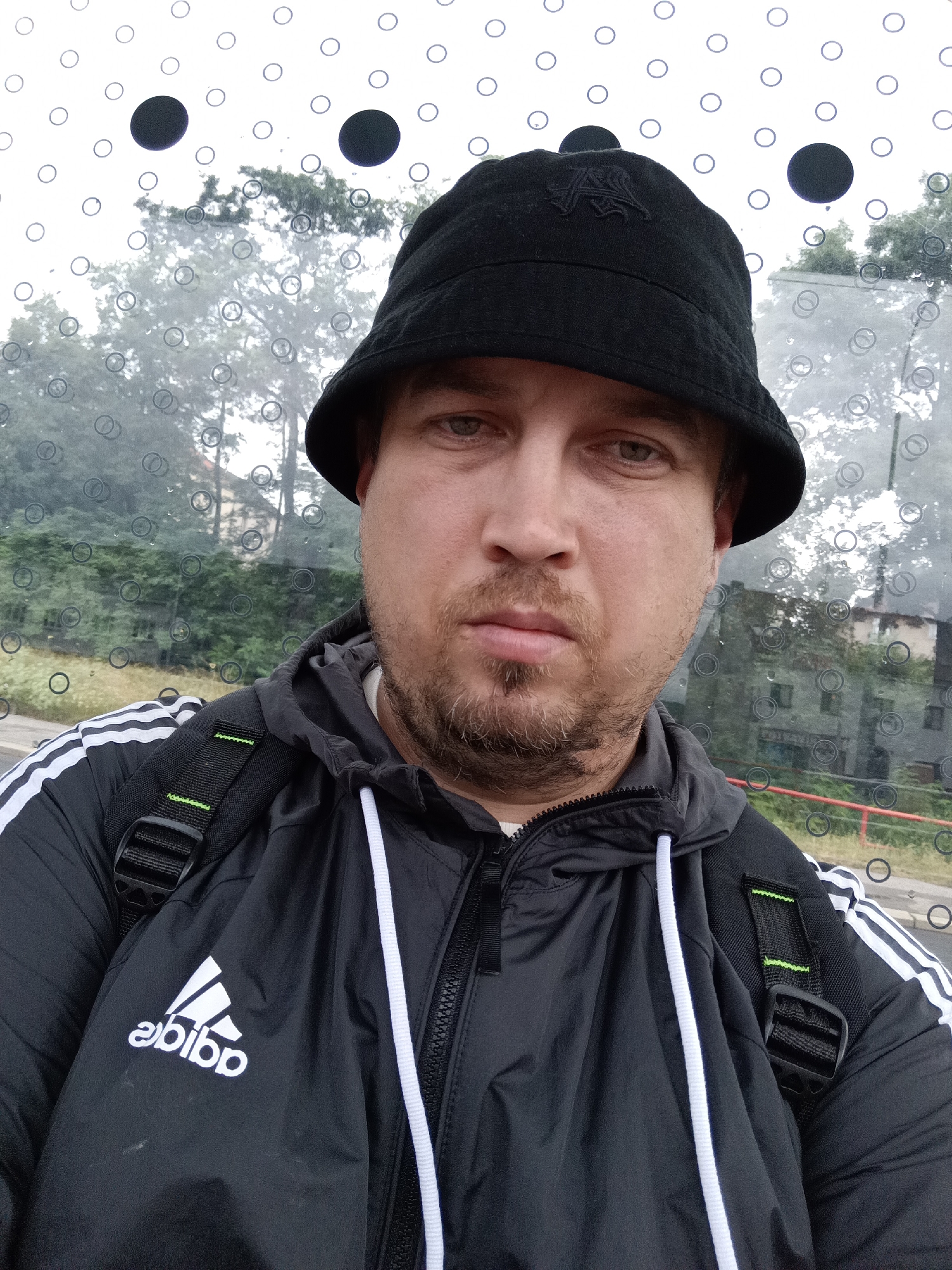 This is a selfie of a man wearing a black bucket hat and a black Adidas jacket with white stripes on the sleeves. He has a short beard and is looking directly at the camera with a neutral expression. He is wearing a backpack. The background appears to be a window with circular patterns and reflections. Outside the window, trees and buildings are visible, suggesting an outdoor setting. The lighting suggests it might be daytime, possibly overcast. The presence of buildings and foliage indicates an urban or suburban environment. There is no specific activity depicted, it appears to be a posed self-portrait. The Adidas logo is clearly visible on the jacket. The circular patterns on the window might be a design feature of a bus stop or a similar public shelter, which, combined with the outdoor scenery, suggests the man is waiting for public transportation or is in a transit area.