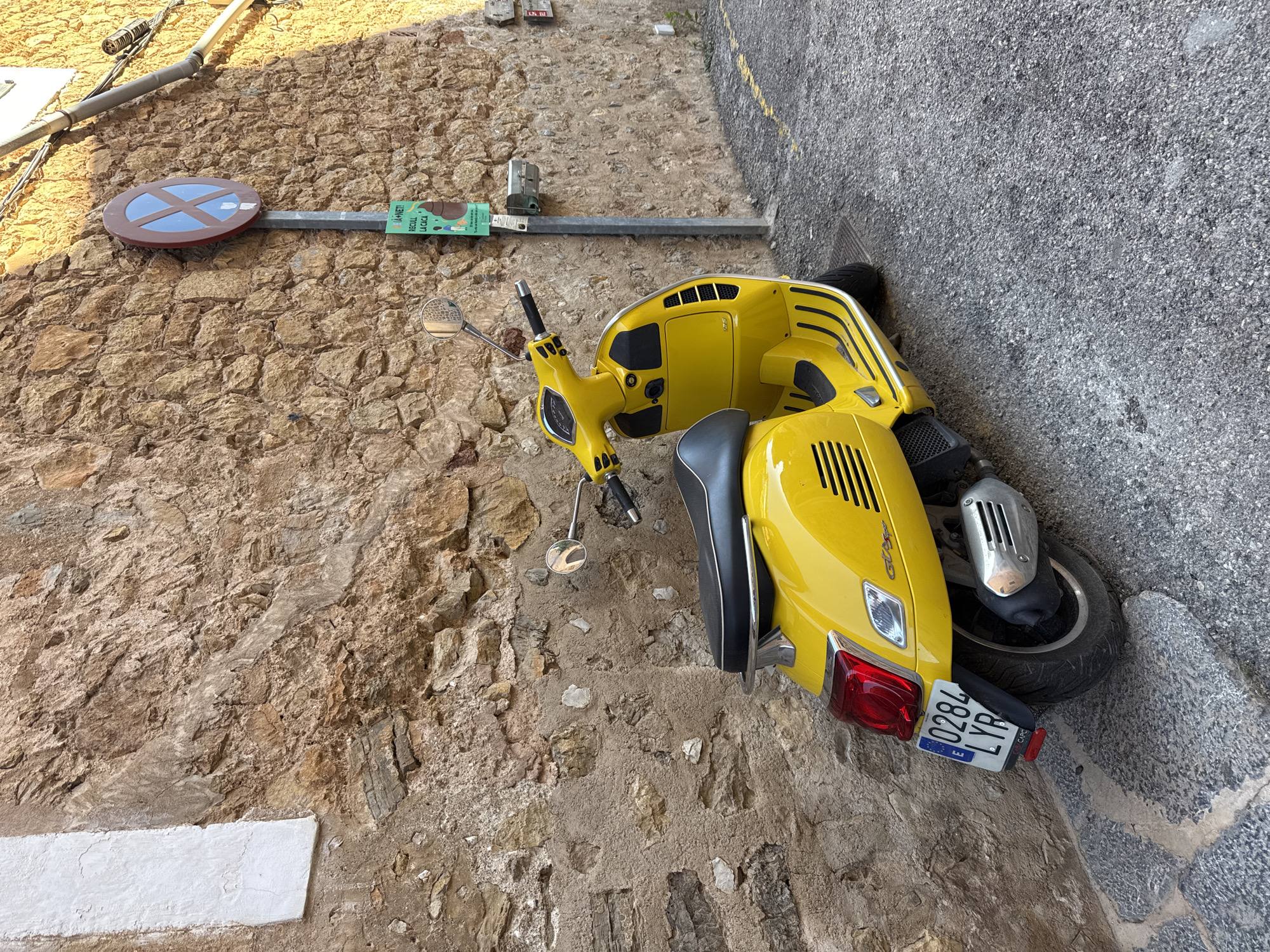 A yellow Vespa scooter is parked on a stone-paved surface against a stone wall in Deià, Spain. The scooter is positioned at an angle to the viewer with the rear of the vehicle closest to the wall. The rear license plate reads "0284 LYP." A circular blue and brown street sign on a metal pole, a small green and white rectangular sign, and electrical conduit are mounted on the stone wall. The image appears to be taken from above, looking down. A pair of shoes sits at the top of the image.
