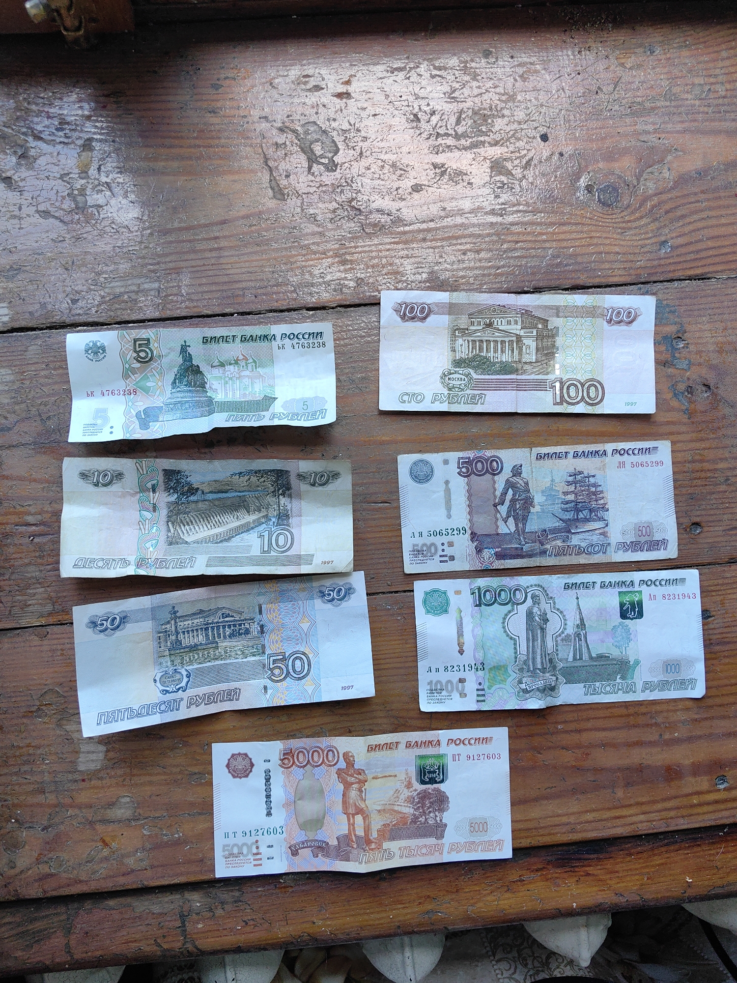 The image displays a collection of Russian rubles laid out on a weathered wooden surface. The bills are arranged in various positions, some overlapping. The denominations visible are 5, 10, 50, 100, 500, 1000, and 5000 rubles. Each bill features distinct imagery and text, including the denomination, the words "БИЛЕТ БАНКА РОССИИ" (Bank of Russia Ticket), and serial numbers. Some of the images on the bills include architectural landmarks like the Bolshoi Theatre in Moscow, the monument to Peter the Great in St. Petersburg, and other historical or cultural depictions. The wooden surface appears rustic, with visible grain and some signs of wear. There are no people or any indication of weather or time of day. The scene is solely focused on the currency.
