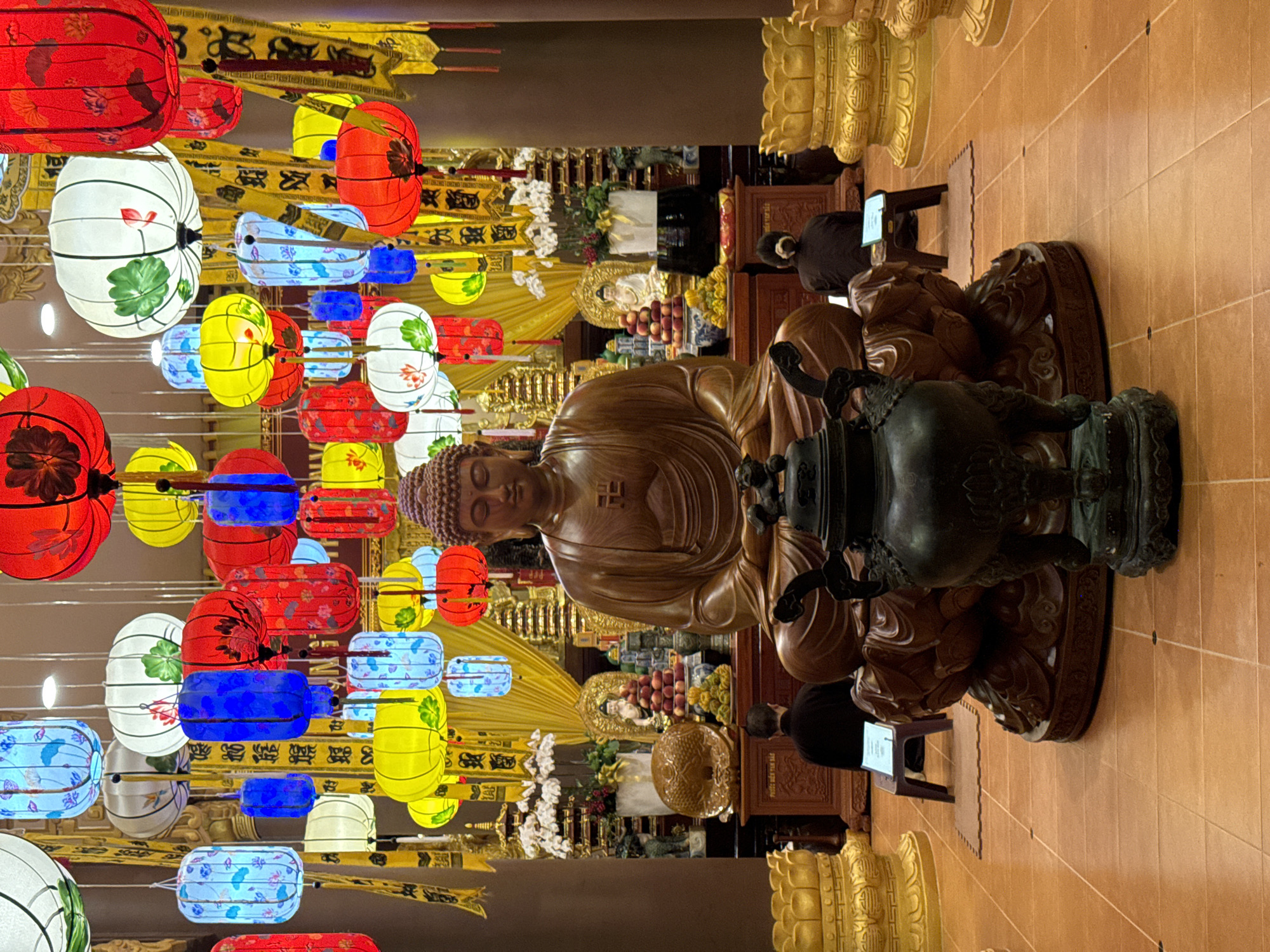 The image shows the interior of the Thích Ca Phật Đài temple in Mũi Né, Vietnam. A wooden Buddha statue sits in the center, with a black incense burner in front of it. People are praying in the background. Colorful lanterns hang overhead. The floor is tiled. Golden statues adorn the periphery of the scene.