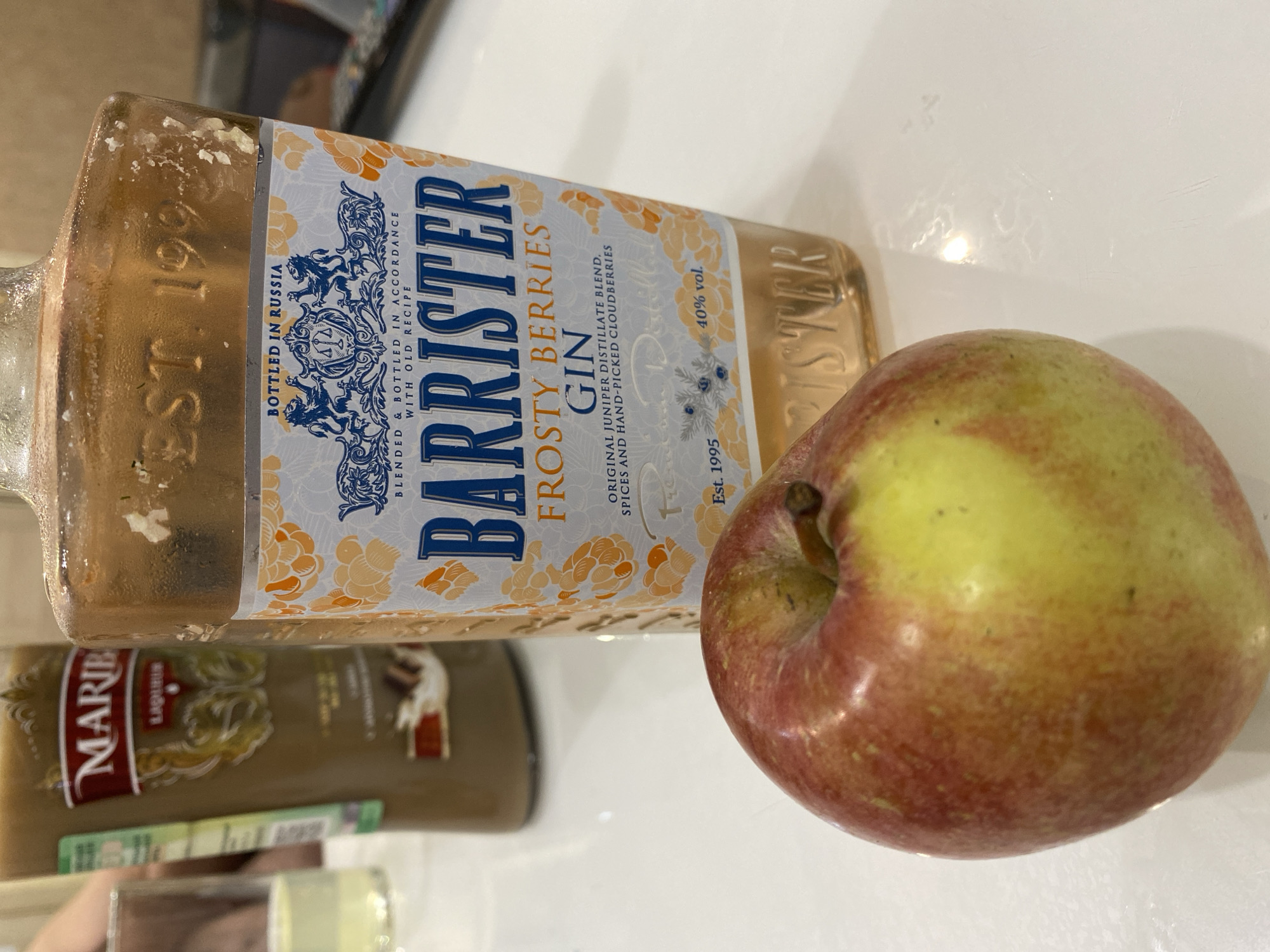 The scene is located in Tyumen, Russia. The foreground features a red and yellow apple with its stem visible, positioned on a clean, white, reflective surface. Behind the apple, a clear glass bottle of "BARRISTER FROSTY BERRIES GIN" stands prominently. The gin bottle's label, featuring a blue and white design with a coat of arms displaying two lions and a scale, states "BOTTLED IN RUSSIA" and specifies "BLENDED & BOTTLED IN ACCORDANCE WITH OLD RECIPE" using "ORIGINAL JUNIPER DISTILLATE BLEND, SPICES AND HAND-PICKED CLOUDBERRIES." The alcohol content is listed as "40% vol." The bottle also indicates "Est. 1995" both on the label and embossed into the glass near the top, which shows some white, crystal-like residue.
In the blurred background to the left, a tall, dark brown bottle with a visible label reading "MARIB LIQUIR" is partially seen. Further left, a clear glass contains a pale yellow liquid. A person's arm or shoulder is faintly discernible in the far left background, indicating human presence. The overall setting appears to be an indoor environment, likely a kitchen or dining area, with consistent bright lighting.