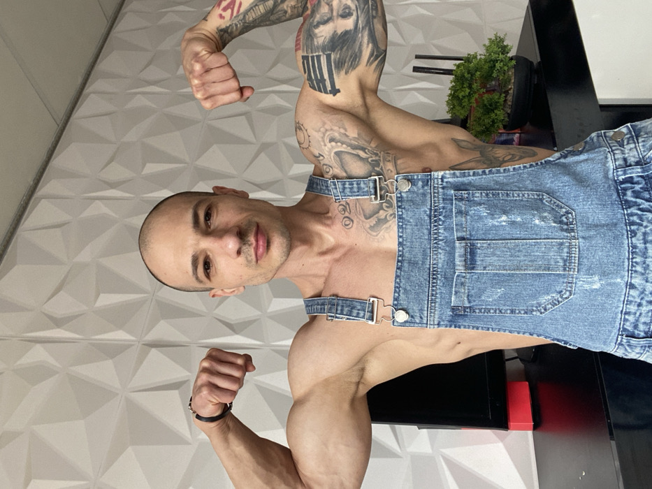 A bald, muscular adult male is depicted indoors, facing the camera with a neutral expression while flexing both biceps, fists clenched. He wears distressed light blue denim overalls over a bare torso, revealing extensive tattoos across both arms and shoulders, with a black bracelet on his left wrist. The background features a white wall with a geometric three-dimensional pattern. To the subject's left, a dark flat-screen monitor and a red rectangular object rest on a dark surface. To his right, a small potted green plant is visible on the same surface. No visual markers within the image indicate a specific location in Pleven, Bulgaria.