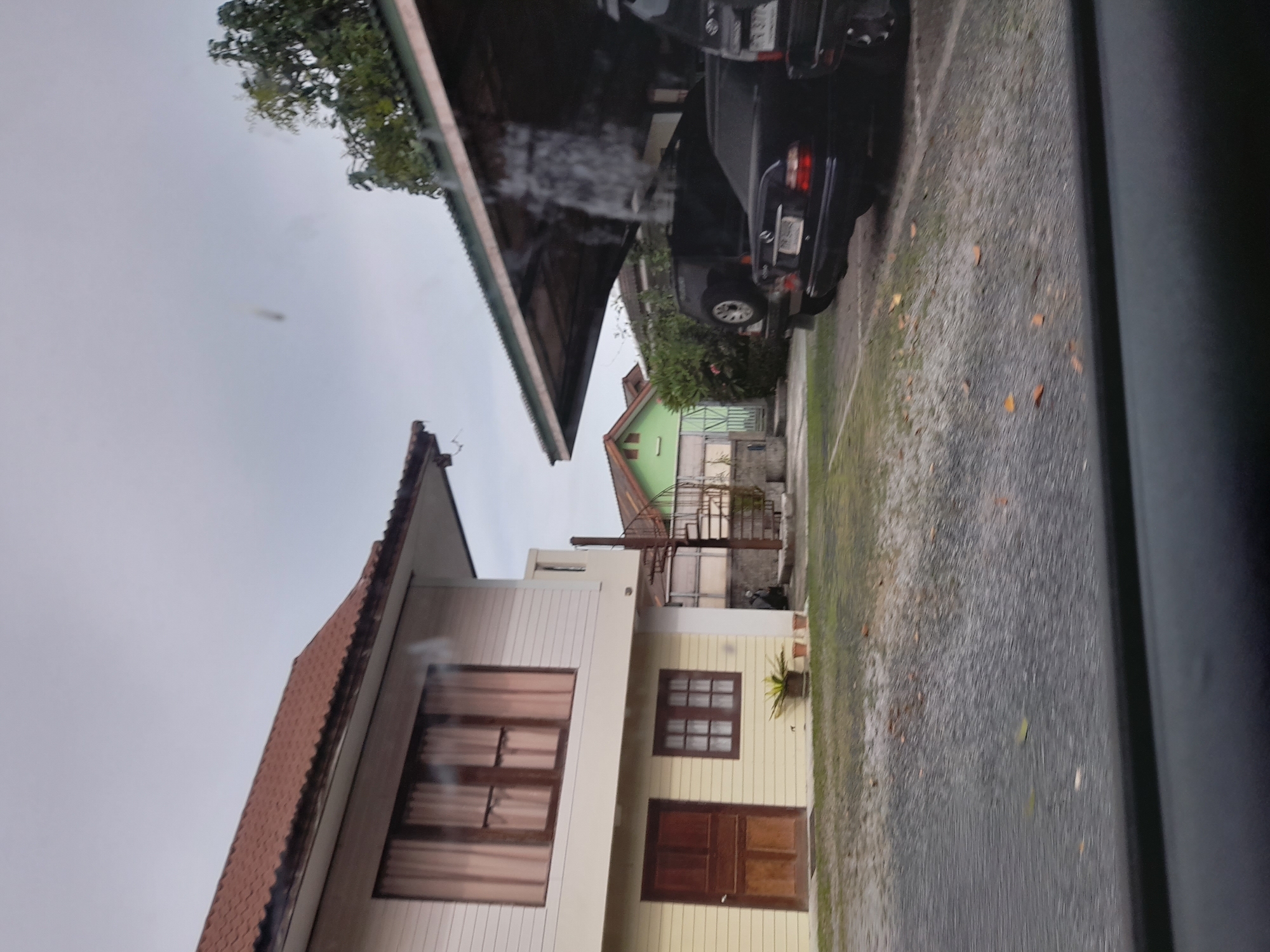 The image depicts a residential area in Baan Mae Nam, Thailand, under an overcast sky. The foreground features a gray, gravel-covered driveway with scattered brown leaves. A dark-colored car is parked on the right, partially obscured by a covered structure with a corrugated metal roof. To the left, a two-story house with white siding on the upper floor and yellow siding on the lower floor is visible. The house has multiple windows with brown frames and sheer curtains, and a dark brown wooden door on the ground level. A spiral staircase, appearing to be made of metal, is attached to the side of the house. In the background, another house with a green exterior and an orange roof is situated behind a low wall and some greenery. The overall impression is of a quiet, somewhat weathered, residential setting.