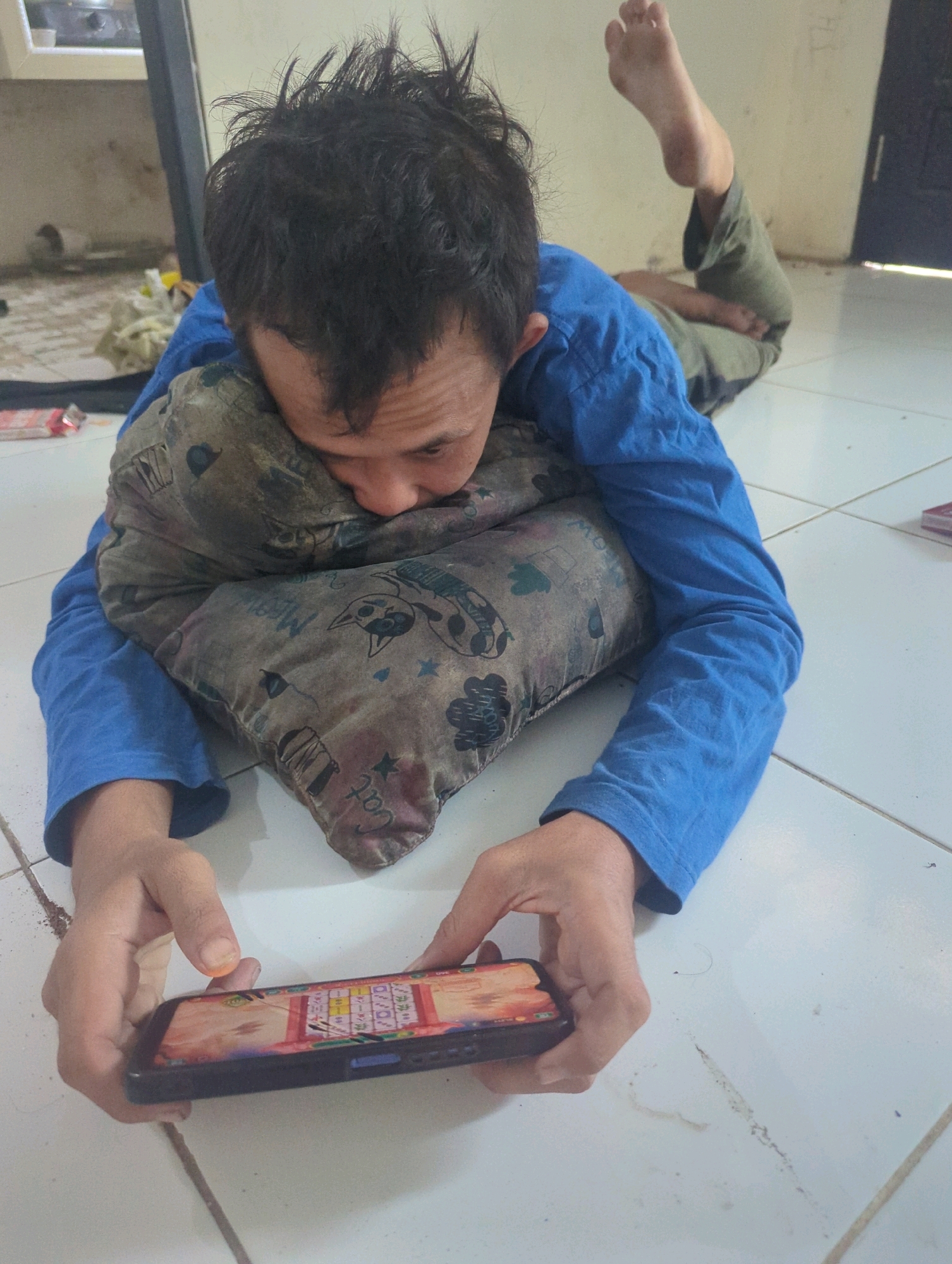 A person is lying on a tiled floor, resting their head on a patterned pillow. They are wearing a blue long-sleeved shirt and olive green pants. Their feet are propped up in the air. The person is holding a smartphone in their hands and appears to be looking at the screen. The phone displays a game with a grid of colorful tiles. The environment seems to be indoors, with part of a wall visible in the background. The floor has some dirt and dust. There are some items on the floor in the background, including what appears to be a box or packaging and some fabric. The overall scene suggests a casual and relaxed setting. There is no specific indication of the location being in Batu Belaman, Indonesia, beyond the prompt's directive.