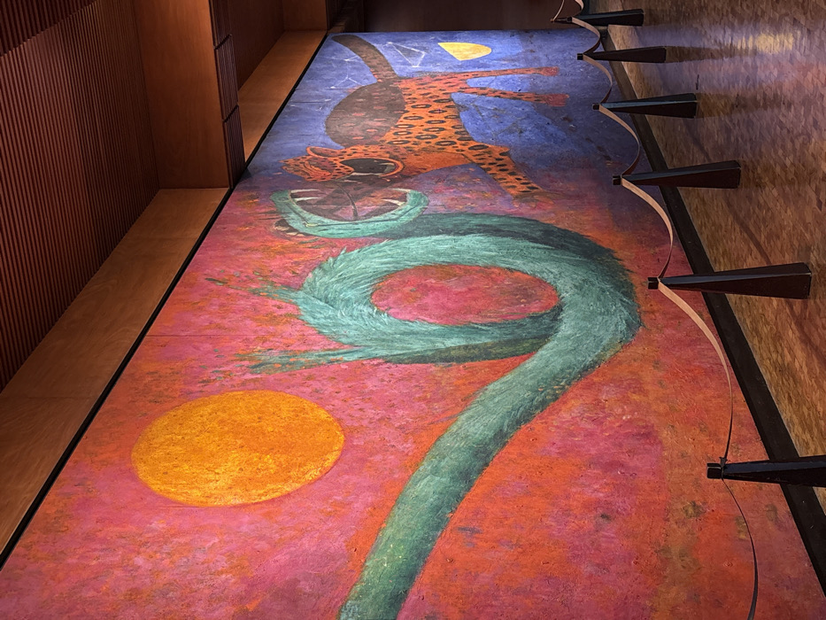 This image depicts a large mural titled "Dualidad" (Duality) by Rufino Tamayo, located on a wall within the Museo Nacional de Antropología (National Museum of Anthropology) in Chapultepec Park, Mexico City, Mexico.
The mural itself is a vibrant, textured painting occupying a significant portion of the wall. The left side of the mural features a dominant red and orange background, against which a large, circular orange form, representing the sun, is positioned in the upper left. A large, turquoise-green feathered serpent-like creature with an open mouth and fangs emerges from the lower left, coiling upwards and to the right. The right side of the mural transitions into a deep blue and purple background. On this side, a reddish-orange jaguar-like feline with black spots, also with an open mouth and visible fangs, is depicted in a lunging posture towards the left. A small yellow crescent, representing the moon, is visible in the lower right corner. Both mythical creatures face each other in an apparent confrontational or dynamic interaction.
The surrounding environment includes a polished, light brown or reddish-brown tiled floor, which reflects light from above. A barrier system, composed of several dark, tapering trapezoidal posts connected by a light-colored rope or strap, runs parallel to the mural in the foreground, indicating a viewing distance. Overhead, a textured wooden ceiling, featuring parallel ribbed panels, extends into the background. No people are visible within the frame.