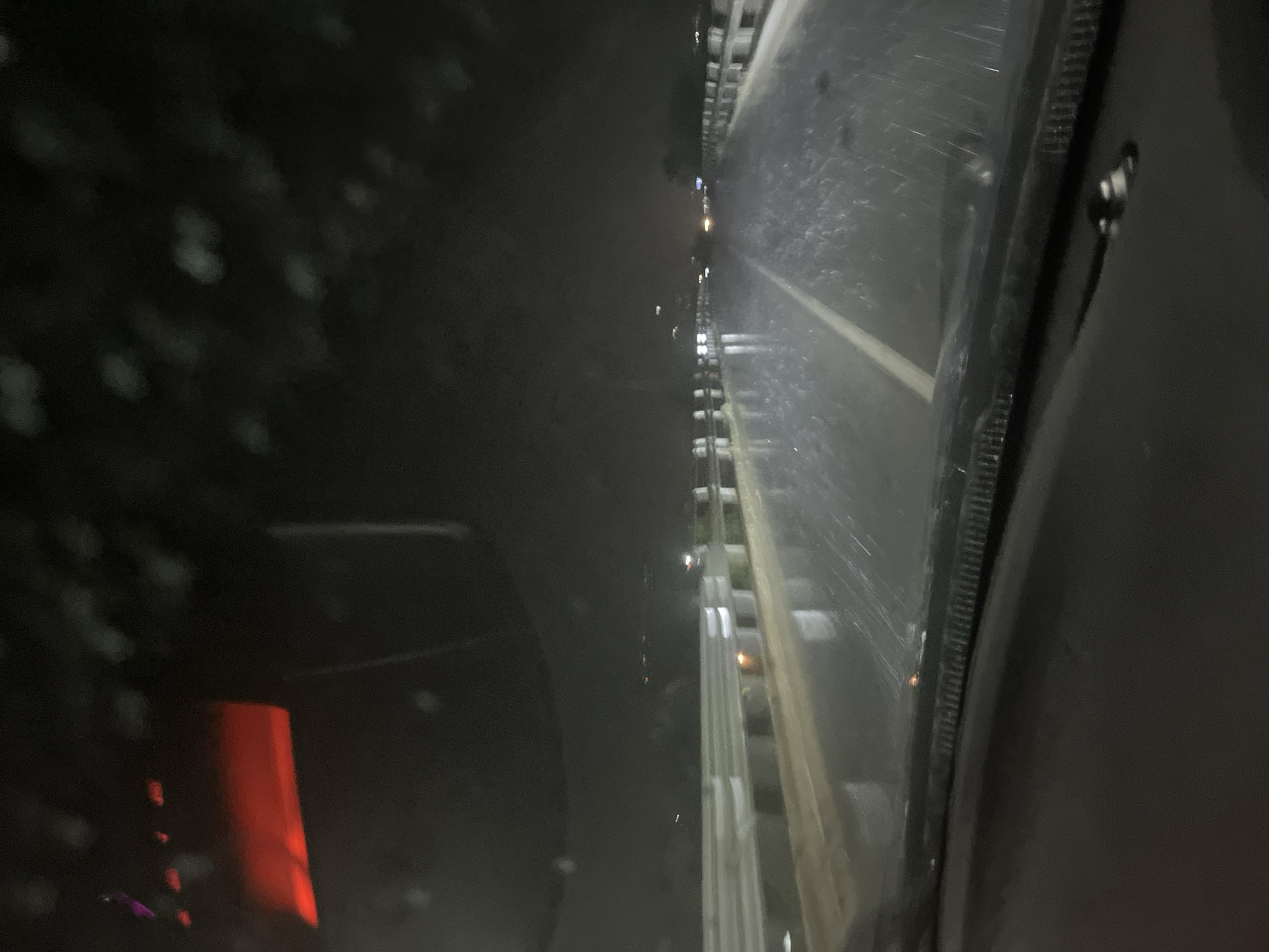 The image captures a nighttime scene from the interior of a vehicle, looking out through a window. The overall illumination is low, relying on artificial light sources.
The left side of the frame is dominated by the dark interior of the vehicle, showing blurry, diffuse light spots that could be reflections on the window's surface or dust particles. In the lower-left portion of this dark area, a prominent, bright red, rectangular object is visible, likely an interior light or part of the vehicle's console.
The right side of the image displays the exterior view through the window. This area reveals what appears to be a road or a linear structure, stretching vertically within the frame. Several artificial lights illuminate this exterior space, appearing as bright points along the structure. A distinct white line, characteristic of a road marking, is visible on the ground surface.
A dark, vertical element, possibly a window frame or a vehicle pillar, acts as a divider between the dark interior on the left and the illuminated exterior view on the right. The image conveys a sense of being in motion or stationary within a vehicle after dark. No people are visible, and there is no discernible text. The conditions appear clear, allowing for visibility of distant lights. The setting is Blagoevgrad, Bulgaria.