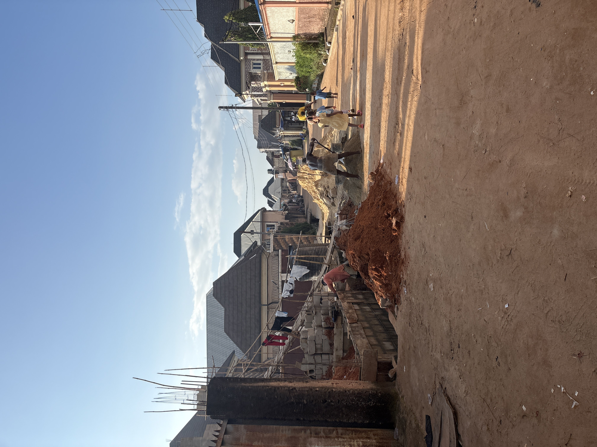 The image depicts a street scene in Obosi, Nigeria, during daylight. The foreground is dominated by a dirt road and construction materials, including piles of red earth and stacks of concrete blocks. On the left side of the road, there is an active construction site with scaffolding, more building materials, and a man working. Laundry hangs on a line above the construction area.
Further down the street, several people are visible. Two children in casual clothing are walking away from the camera, and a man wearing a yellow shirt is also walking in the same direction. Another man, wearing a light-colored shirt and shorts, is pushing a wheelbarrow filled with dirt on the right side of the road, near a large pile of earth.
The background features residential buildings with dark, sloped roofs, typical of the area. Some buildings have decorative facades and walls, suggesting a mix of older and newer constructions. Power lines are strung across the street, supported by utility poles. The sky is clear with a few wispy clouds, and the overall lighting suggests it is either morning or late afternoon. The scene conveys a sense of everyday life and ongoing development in the urban environment of Obosi.