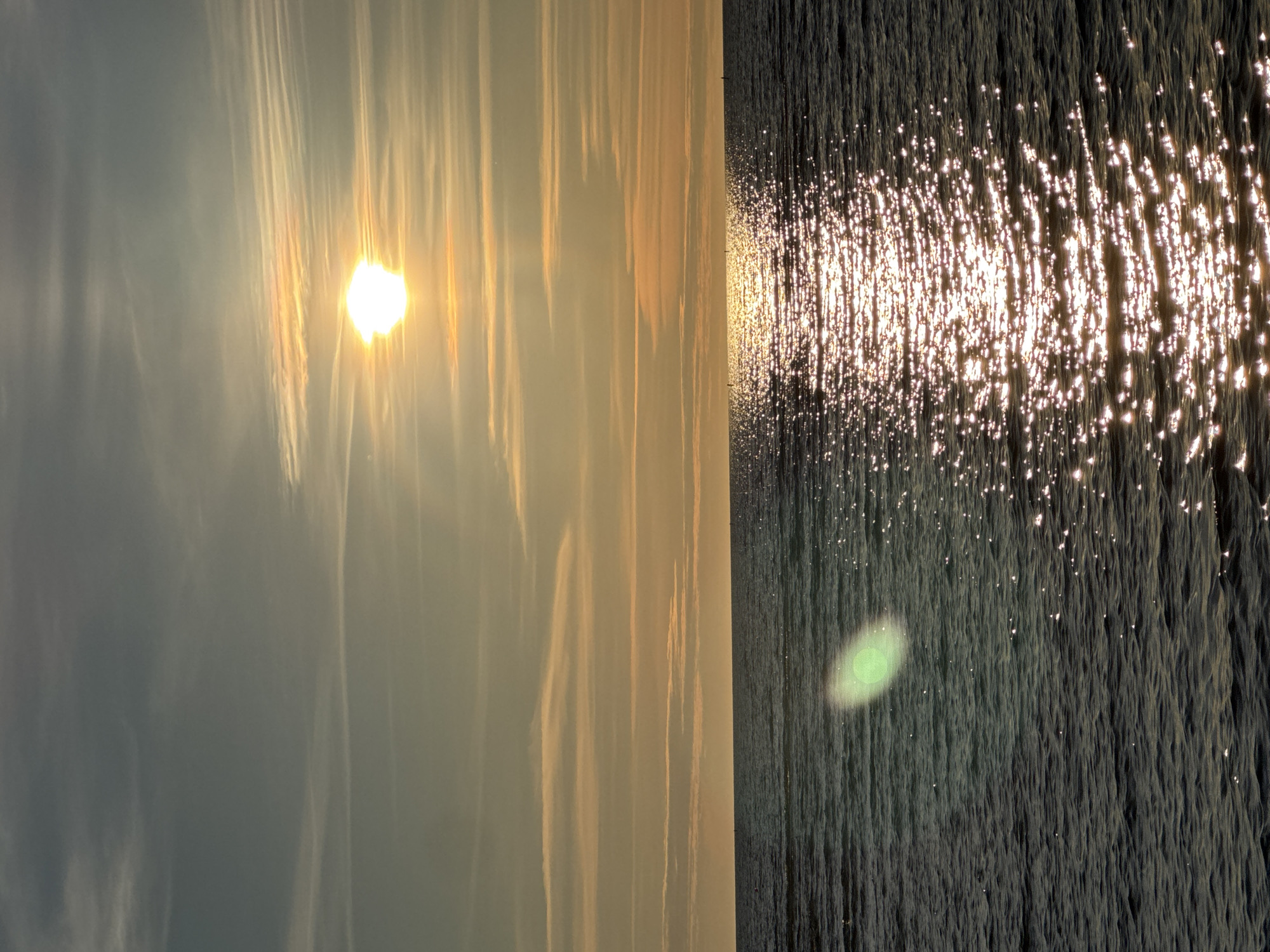 This is a vertical, eye-level shot of the sea at sunset. The sun is a bright, glowing orb in the upper third of the image, casting a shimmering golden path across the dark, rippling water. Wispy, horizontal clouds stretch across the sky, catching the warm light of the setting sun, which appears to be the sun setting over the Baltic Sea, likely near the coast of Tallinn, Estonia. There are no people or man-made structures visible, only the vast expanse of the sea and sky. A faint green lens flare is present on the left side of the water.