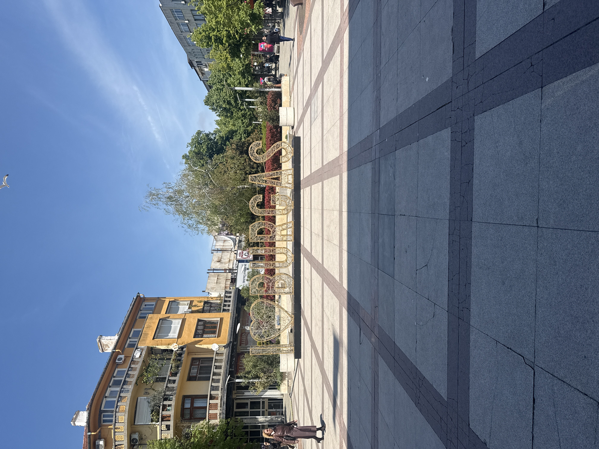 The image depicts a scene in Burgas, Bulgaria. A large decorative sign spelling out "Burgas" in Cyrillic letters, adorned with heart shapes, stands on a paved square. The square is constructed with light gray and darker gray tiles arranged in a grid pattern. To the left of the sign is a multi-story yellow building with multiple windows, balconies, and decorative elements. A woman wearing a dark-colored suit is standing at the bottom right corner of the image. To the right of the sign, trees and a building are seen in the distance. The sky is clear and blue. A seagull is visible in the top left corner of the image.