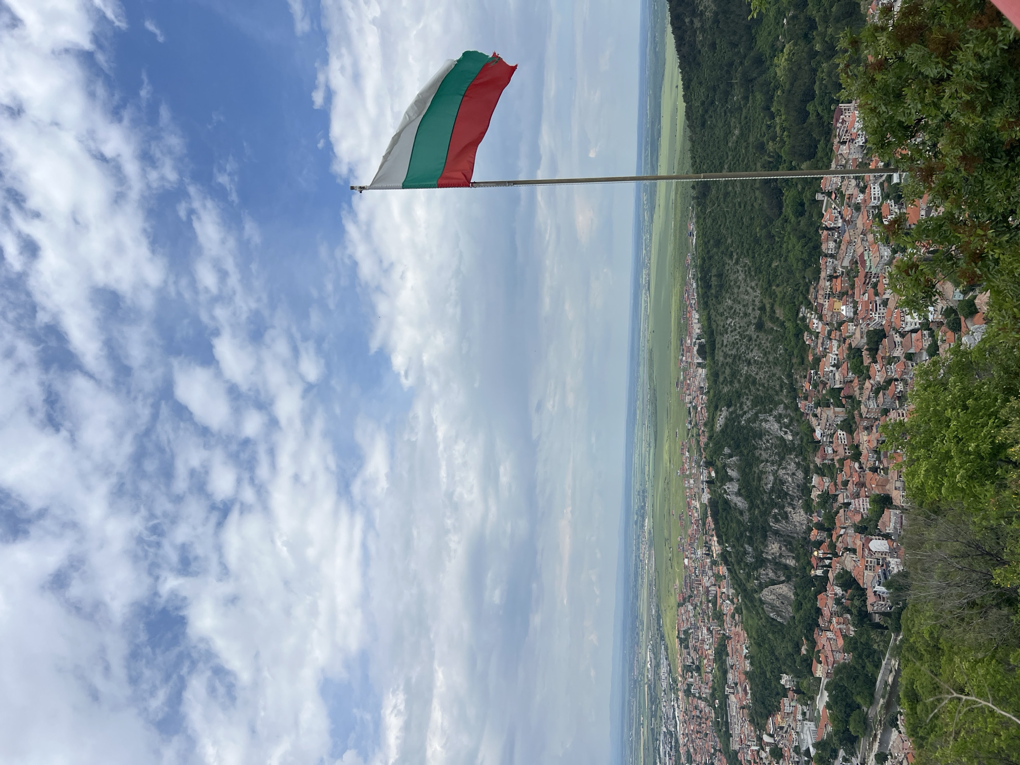 A photograph captures a panoramic view of Asenovgrad, Bulgaria, with the national flag prominently displayed in the foreground. The flag, featuring horizontal stripes of white, green, and red, is extended on a pole against a backdrop of a cloudy sky.
Below the flag, the scene unfolds with a mix of natural and urban elements. A dense forest covers the hillsides, contrasting with the cityscape below. Buildings with red-tiled roofs are clustered together, forming the urban landscape. The terrain slopes downward, leading to a flat expanse of green fields in the distance. The sky above is filled with white clouds against a blue backdrop, contributing to the overall sense of height and perspective.