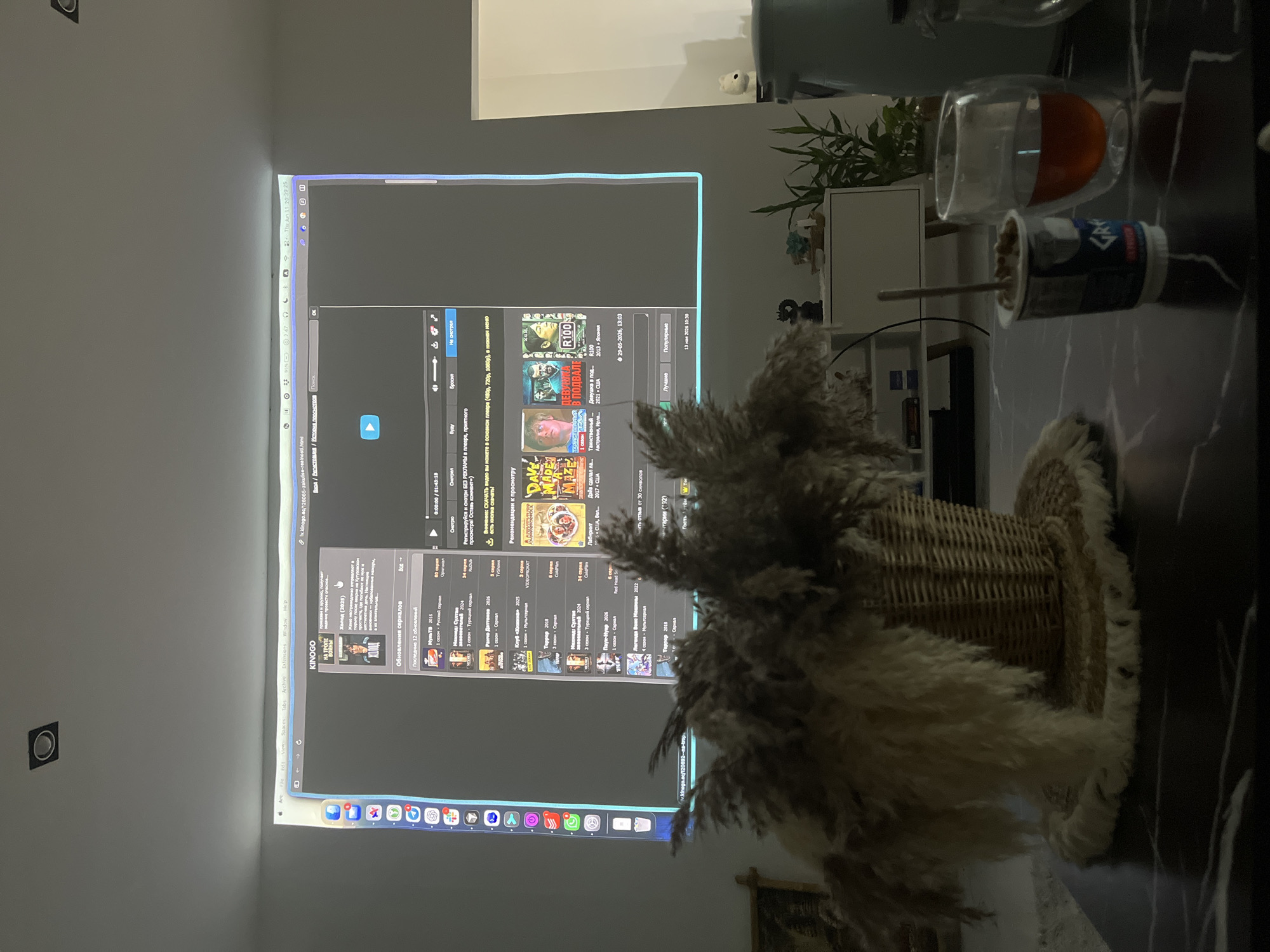 The image displays a projector screen displaying a webpage with various video thumbnails and text in Russian. The webpage appears to be a video streaming or download site. In the foreground, a dark marble table holds a woven basket with dried reeds, a yogurt cup with a spoon, and a glass of amber liquid. The background shows a white furniture unit with a potted plant, and a hint of a white wall with a recessed light fixture at the top of the projected image. The overall scene suggests a relaxed home viewing environment. The location of ตำบลเกาะพะงัน, Thailand is not depicted or suggested by any visual cues in the image.