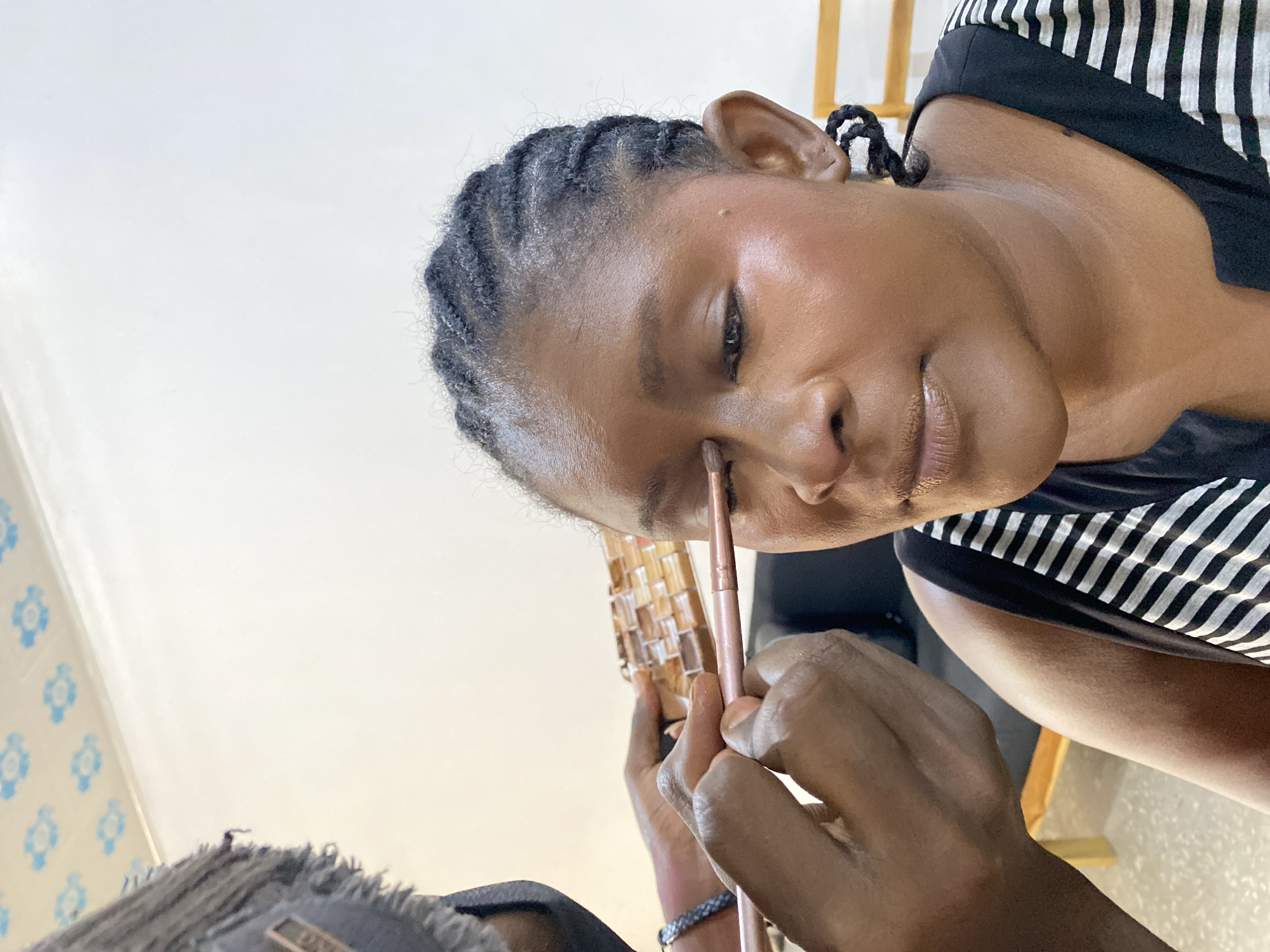 In an indoor setting that appears to be in Gura Top, Nigeria, a woman is receiving makeup application. The woman, with braided hair, is wearing a black and white striped top. A makeup artist is applying makeup to her face with a brush. A makeup palette is visible in the foreground. The background includes a white wall with blue floral patterns and a wooden structure.