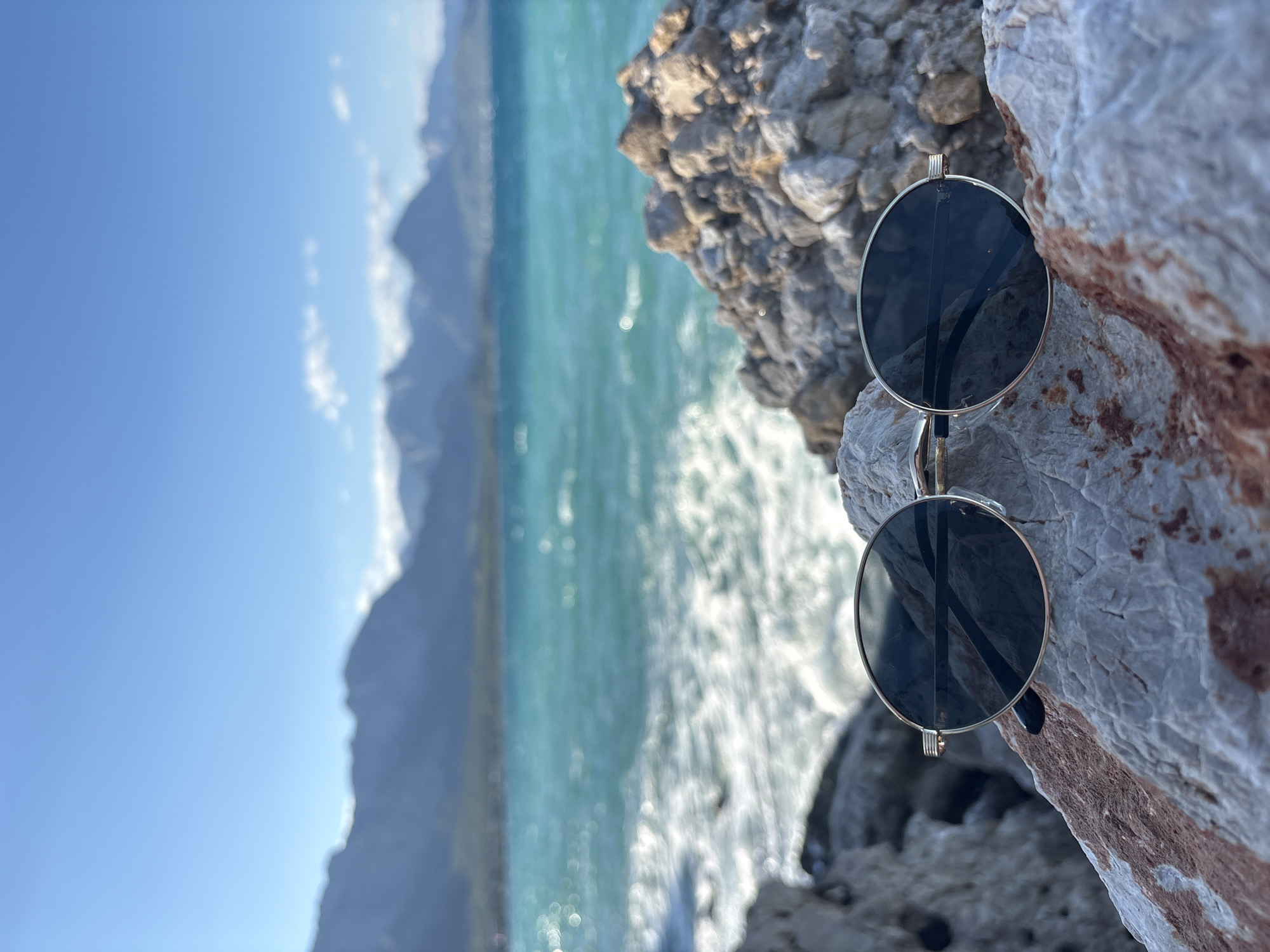 The image captures a pair of round, gold-rimmed sunglasses placed on a rocky surface near the sea. The sunglasses are the central subject, positioned in the foreground and slightly off-center. Behind them, the turquoise waters of the Mediterranean Sea are visible, with waves breaking and creating a foamy white pattern. In the distance, a range of mountains forms the horizon under a clear blue sky. The rocks in the foreground are rough and varied in color, ranging from light gray to reddish-brown. This scene appears to be a coastal area in or near Kemer, Turkey, known for its scenic beaches and mountainous backdrop. There are no people or animals visible in the image.
