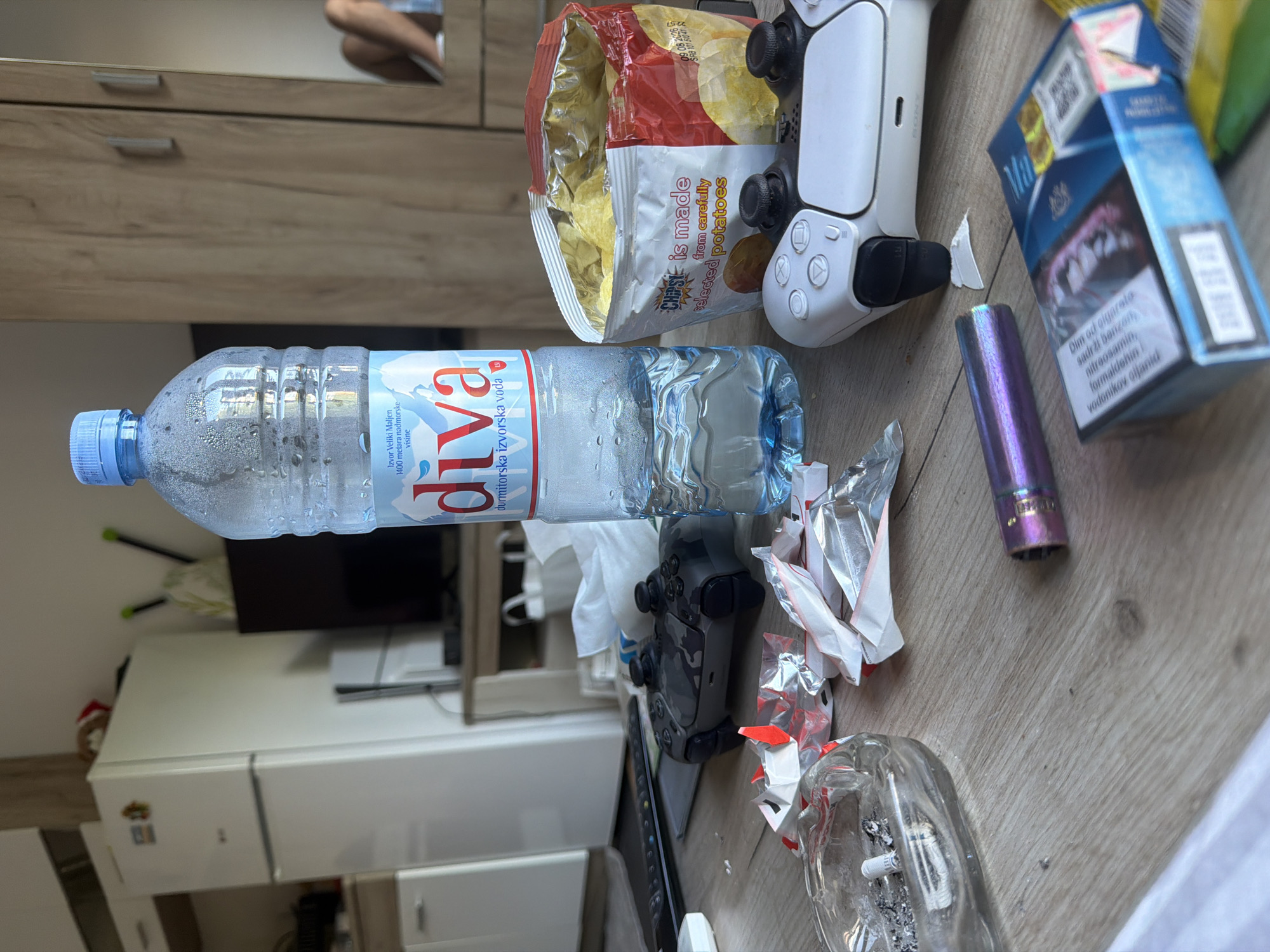 The image depicts a cluttered indoor scene, possibly a living space or bedroom, with various objects scattered across a wooden surface. A partially consumed bottle of "diva" brand water, labelled in Serbian "Izvor Veliki Maljen, 1400 metara nadmorske visine, durmitorska izvorska voda", lies horizontally in the center of the frame. A packet of chips, identified as "Chipsea, is made from carefully selected potatoes" is visible, along with two PlayStation controllers (one white, one black camouflage), and a blue pack of cigarettes. There is also a purple vape pen, crumpled foil packaging, and a glass ashtray filled with cigarette butts.
Based on the product labels (especially the water bottle and cigarette brand), the location is likely somewhere in Montenegro. The city of Podgorica cannot be definitively confirmed from the image alone but is a possibility.