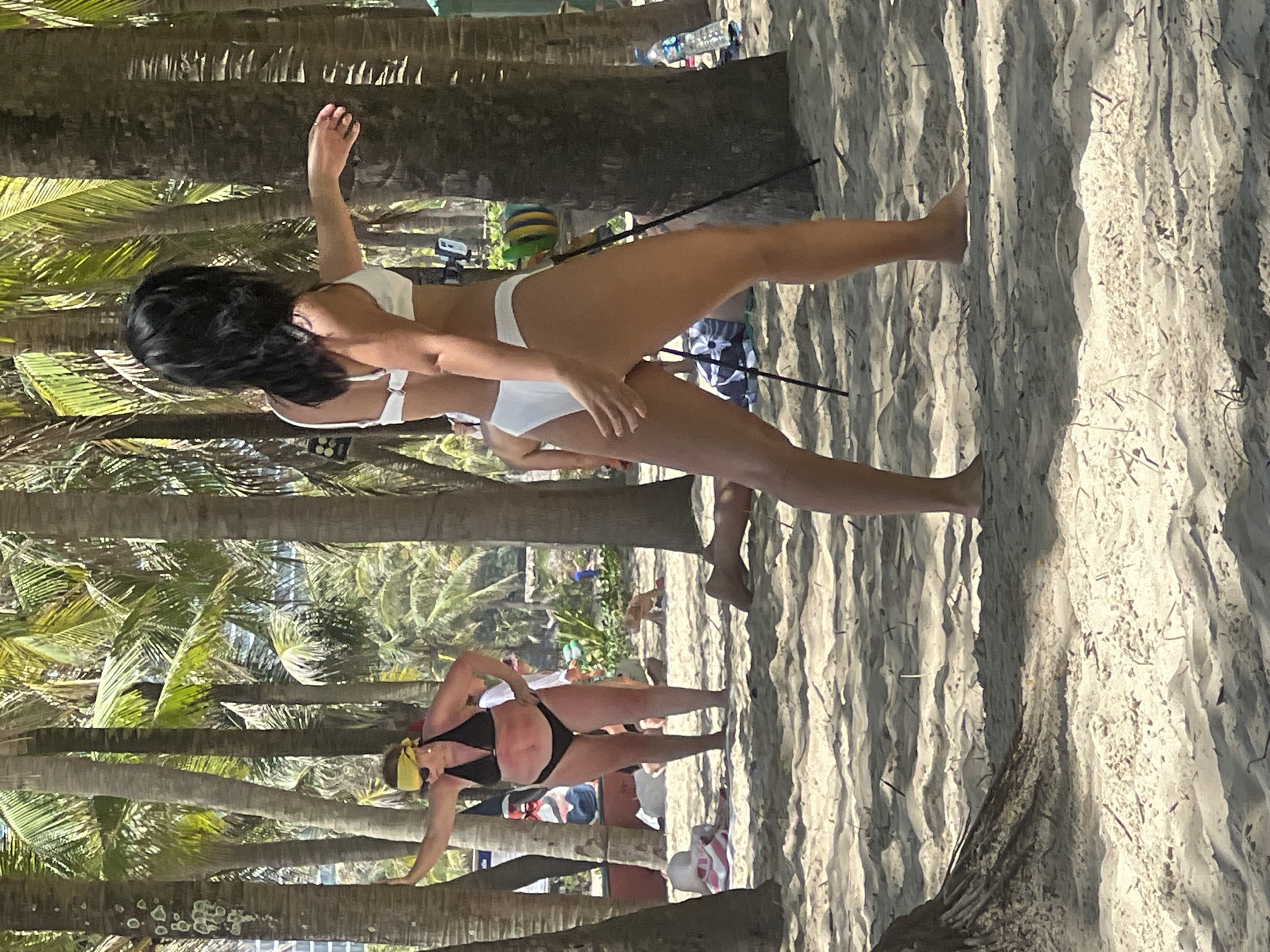The image depicts a sunny beach scene with numerous palm trees. In the foreground, a woman wearing a white bikini is seen from the back, with her right arm extended. To the left, another woman in a black bikini and a yellow bandana is standing with her left arm resting on a palm tree. Both women appear to be engaged in some form of exercise or activity. The sandy beach is scattered with debris and shadows cast by the palm trees. In the background, other people and beachgoers are visible, along with beach umbrellas and a bottle of water. The scene is consistent with a location like My Khe Beach in Da Nang, Vietnam, known for its sandy shores and palm-lined coastline.