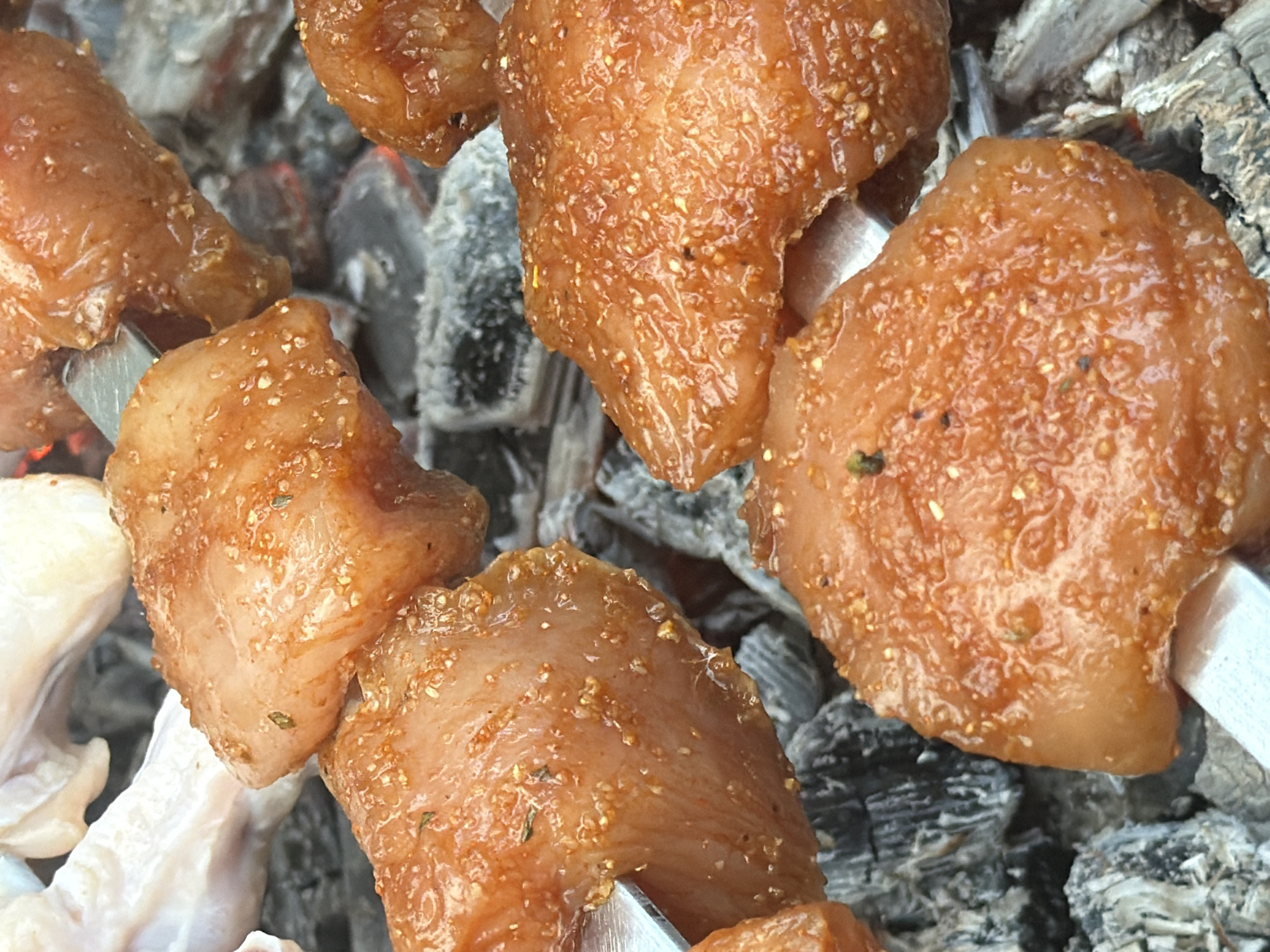This is a close-up, vertical, eye-level, outdoor, slightly overhead shot of raw chicken pieces on metal skewers being cooked over glowing charcoal. The chicken pieces are bite-sized, cubed, and appear to be seasoned with a reddish-brown spice blend that has visible small white specks, possibly salt or sesame seeds. The skewers are inserted into the chicken pieces, with the metal tips visible on some. The chicken is placed directly on a bed of dark grey and black charcoal, some of which are emitting a faint red glow, indicating heat. The background is blurred, but it appears to be more charcoal and possibly some partially cooked chicken or other food items in the upper left corner, which are lighter in color and appear to be chicken wings. The focus is sharp on the chicken pieces and the charcoal. There are no people or other discernible objects in the scene. This scene depicts food preparation over a grill. The location in the city of Tuapse, Russia is not evident from the image itself.