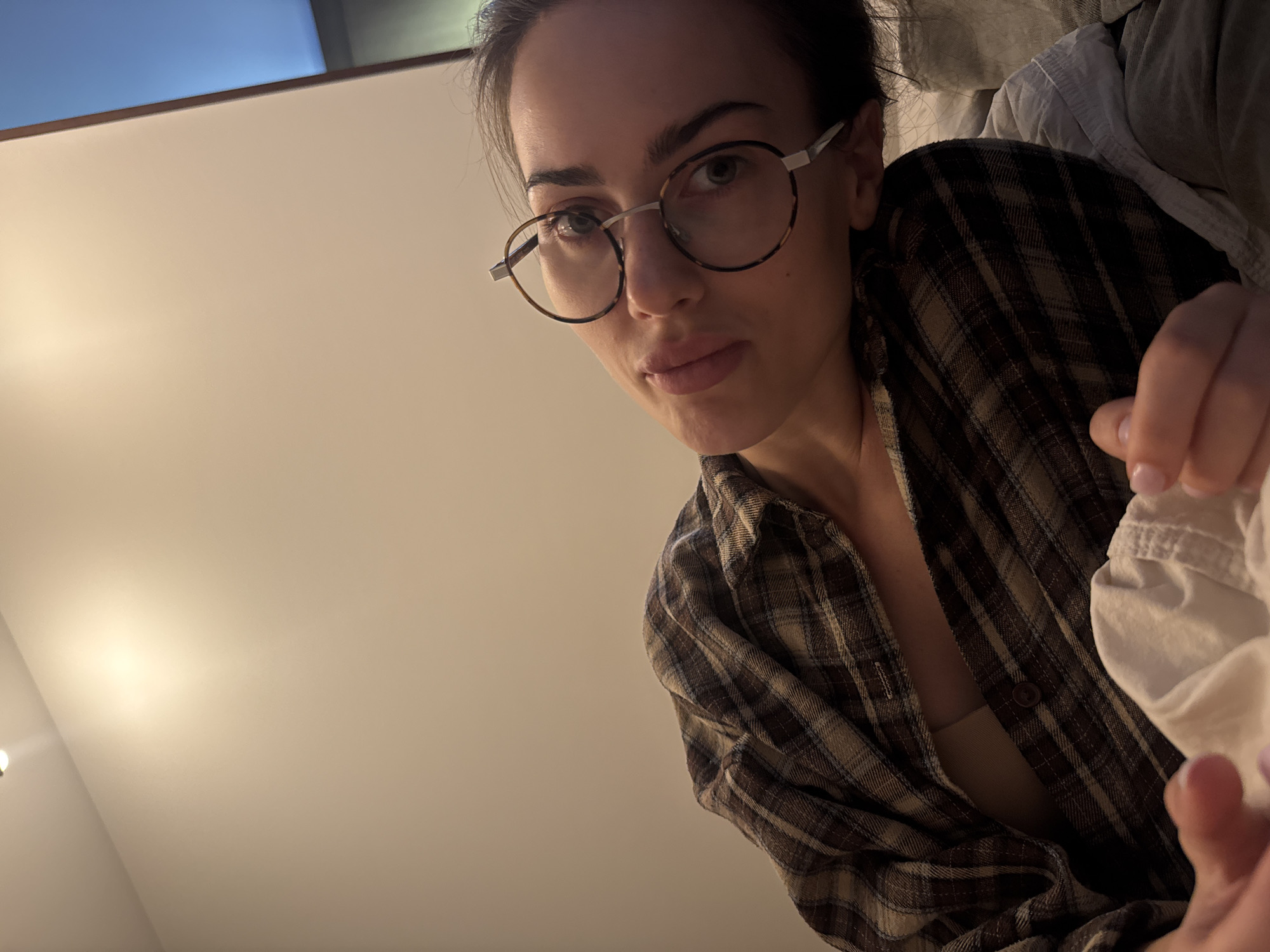 A woman with dark hair, fair skin, and wearing round glasses is seen from a low angle perspective. She is wearing a brown and tan plaid shirt. The background is a plain off-white ceiling with a slight blue tint in the upper left corner. The location is unknown.