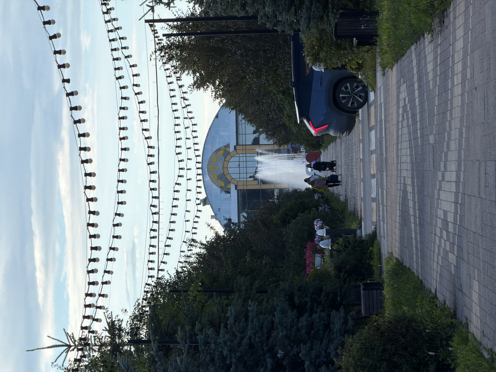 A group of people are walking down a paved walkway beside a fountain. The walkway is lined with trees and bushes, and there are decorative lights strung overhead. In the background, a large building with a curved facade and arched windows can be seen. A dark-colored car is parked on the side of the walkway, with its taillights illuminated. The scene appears to be set in an urban park or plaza. The lighting suggests it might be late afternoon or early evening.