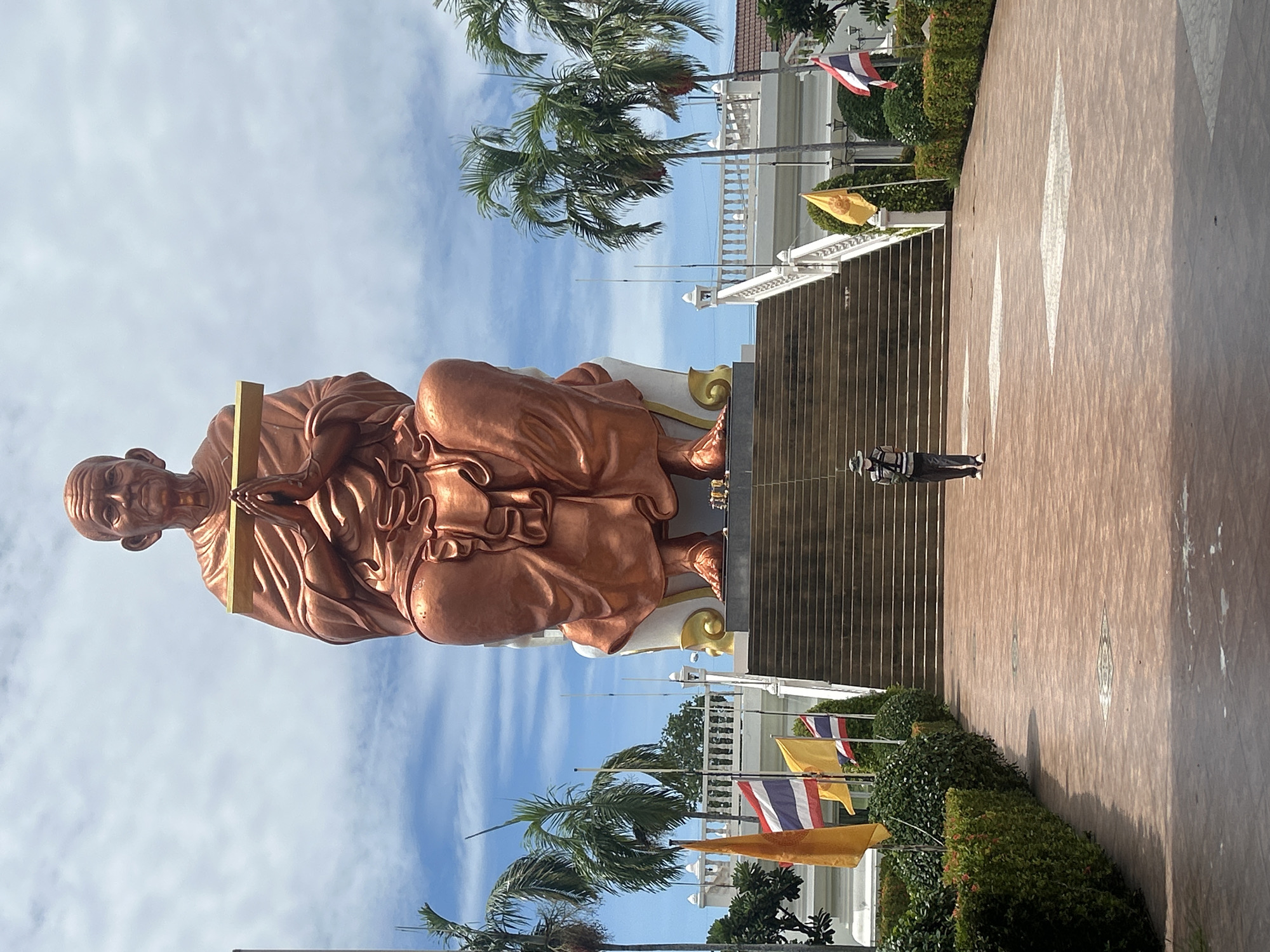 A full shot captures a colossal bronze statue of a seated Buddhist monk, hands pressed together in a prayer gesture, holding a golden staff. The statue is perched atop a grand staircase, flanked by lush green hedges and tall palm trees. Several flags, including the Thai flag, are visible. A lone individual, wearing a hat and vest, stands on the paved walkway leading towards the stairs, gazing up at the imposing figure. The sky above is partly cloudy, suggesting a bright day. The location is identified as Thai Ko Subdistrict, Thailand.