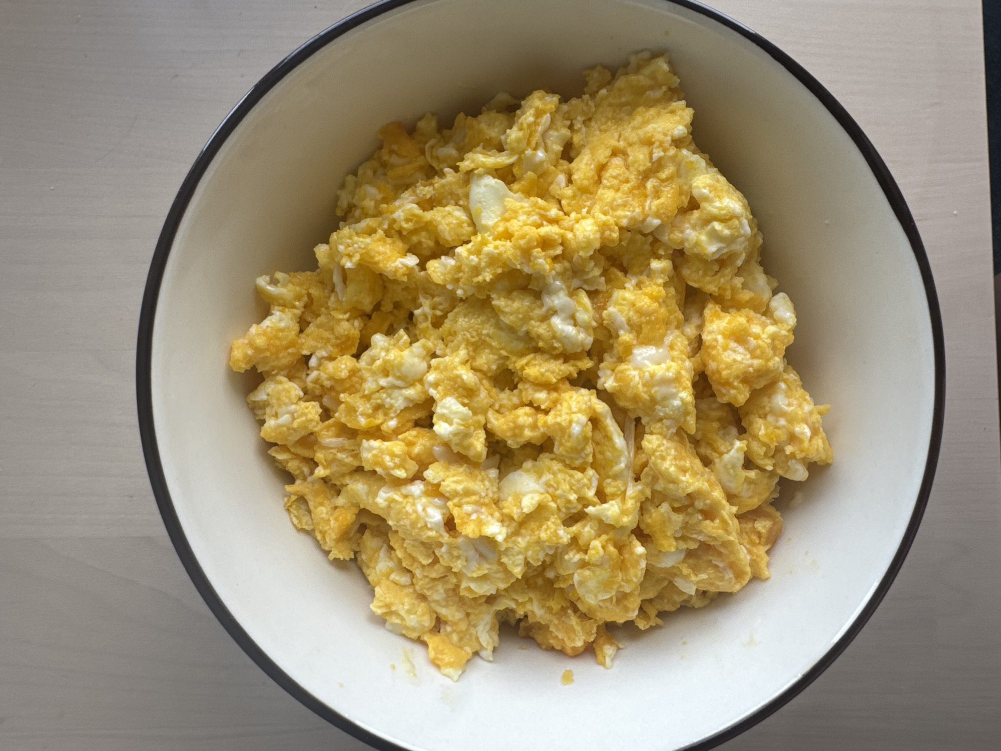 A bowl of fluffy, yellow scrambled eggs sits on a light brown wooden table. The eggs fill most of the off-white bowl, which has a dark rim. The lighting suggests a natural, overhead light source. No people or other objects are visible in the image. The scene is indoors. The image was taken on the 11th of April, 2024.