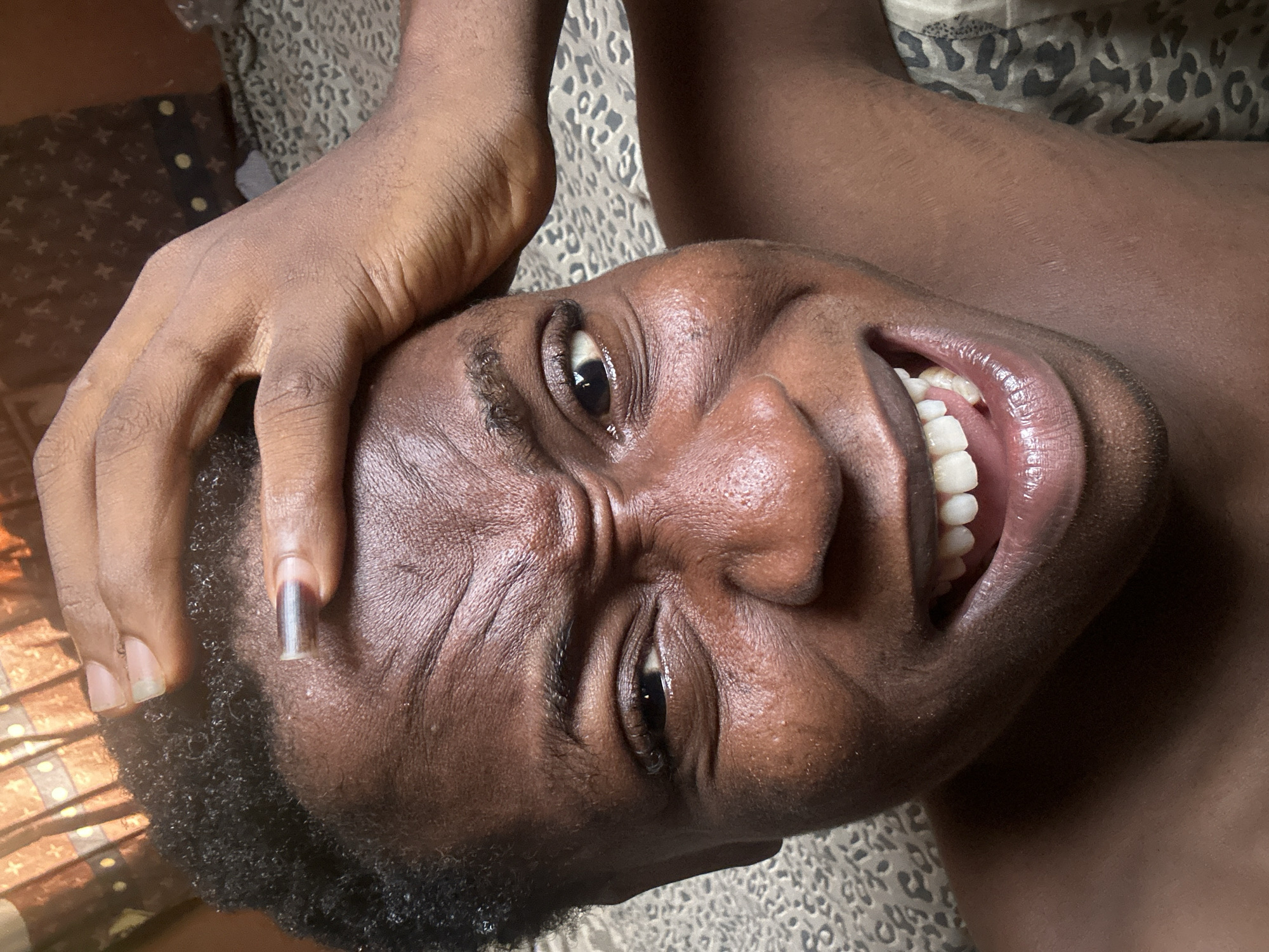 The image is a close-up of a person's face, lying down. The person is smiling widely, revealing their teeth. Their hand is on their head. The background consists of a patterned surface, possibly a bedspread or blanket. There is nothing in the image to indicate a specific location in a city.