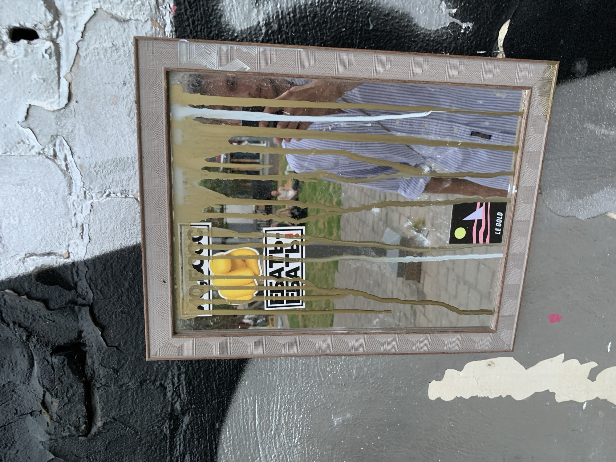 A framed mirror, approximately 18 inches by 14 inches, hangs on a heavily textured wall in Tel Aviv, Israel. The wall is a combination of peeling white paint, black graffiti, and grey concrete. Gold and white paint streaks vertically down the mirror's surface. Stickers adhere to the mirror's surface including one featuring a yellow lemon and another reading "LE GOLD." The mirror reflects a portion of the street, a striped shirt, and a human face. The mirror's frame is patterned with a light grey geometric design.