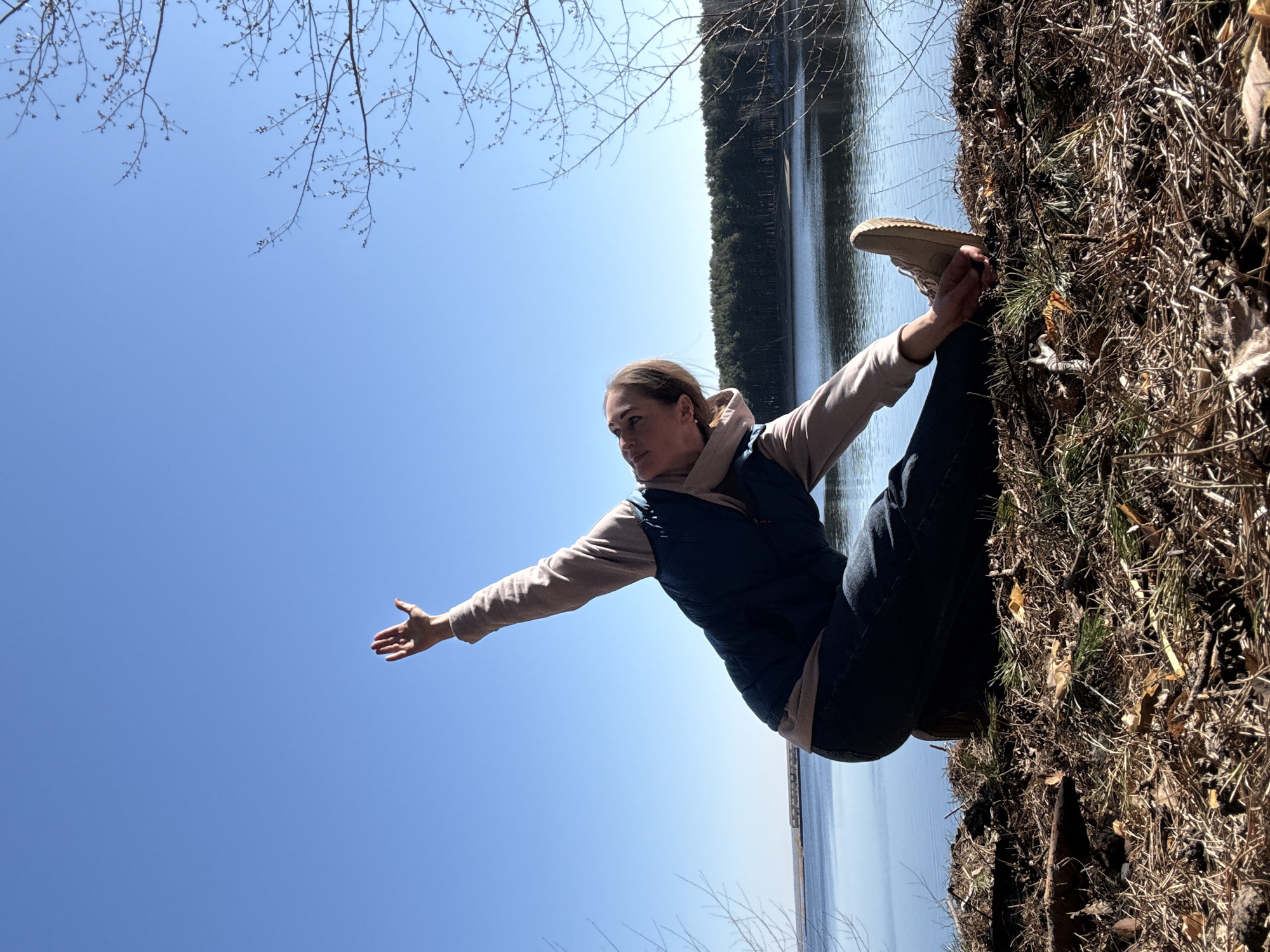 A woman is performing a yoga pose near a body of water. She is wearing dark-wash jeans, a beige hooded sweatshirt, and a navy blue vest. Her right arm is extended towards the clear blue sky. She is holding her left foot with her left hand. The background consists of trees and a body of water, possibly a lake or river, suggesting a natural setting near the city of Chaikovsky, Russia. The ground near her is covered with dry grass and leaves. The photo is taken from a low angle and rotated.