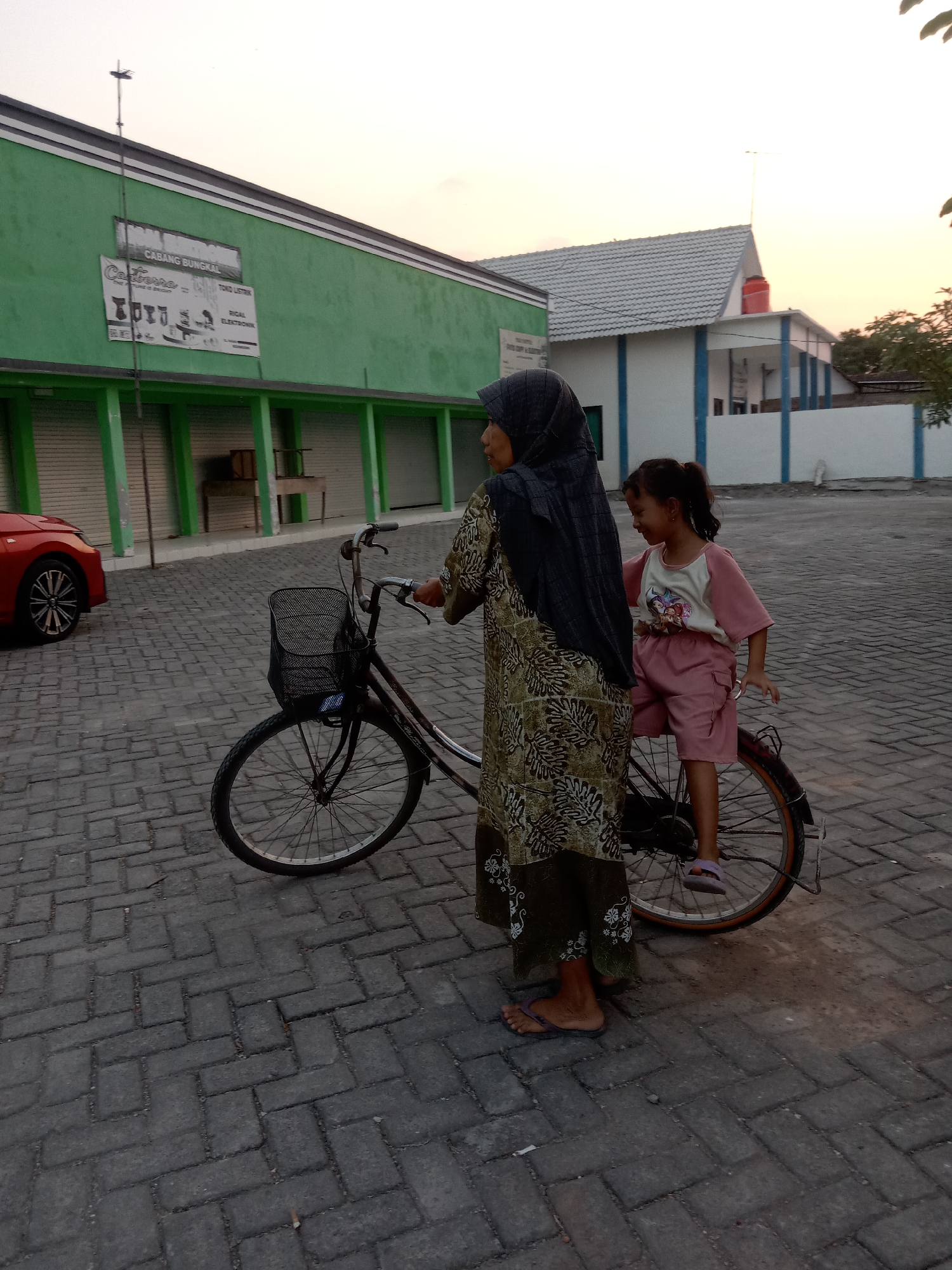In the city of Stiaji, Indonesia, a woman wearing a hijab and a long, patterned dress stands next to a bicycle with a basket. A young girl in a pink shirt and shorts sits on the back of the bicycle, looking to her right and smiling. The bicycle is dark in color, with a wire basket on the front. The ground is paved with grey bricks. In the background, there is a long, green building with rolling shutters, and a white building with a grey tiled roof. A red car is parked to the left of the frame. The sky is a pale orange, indicating either sunrise or sunset.