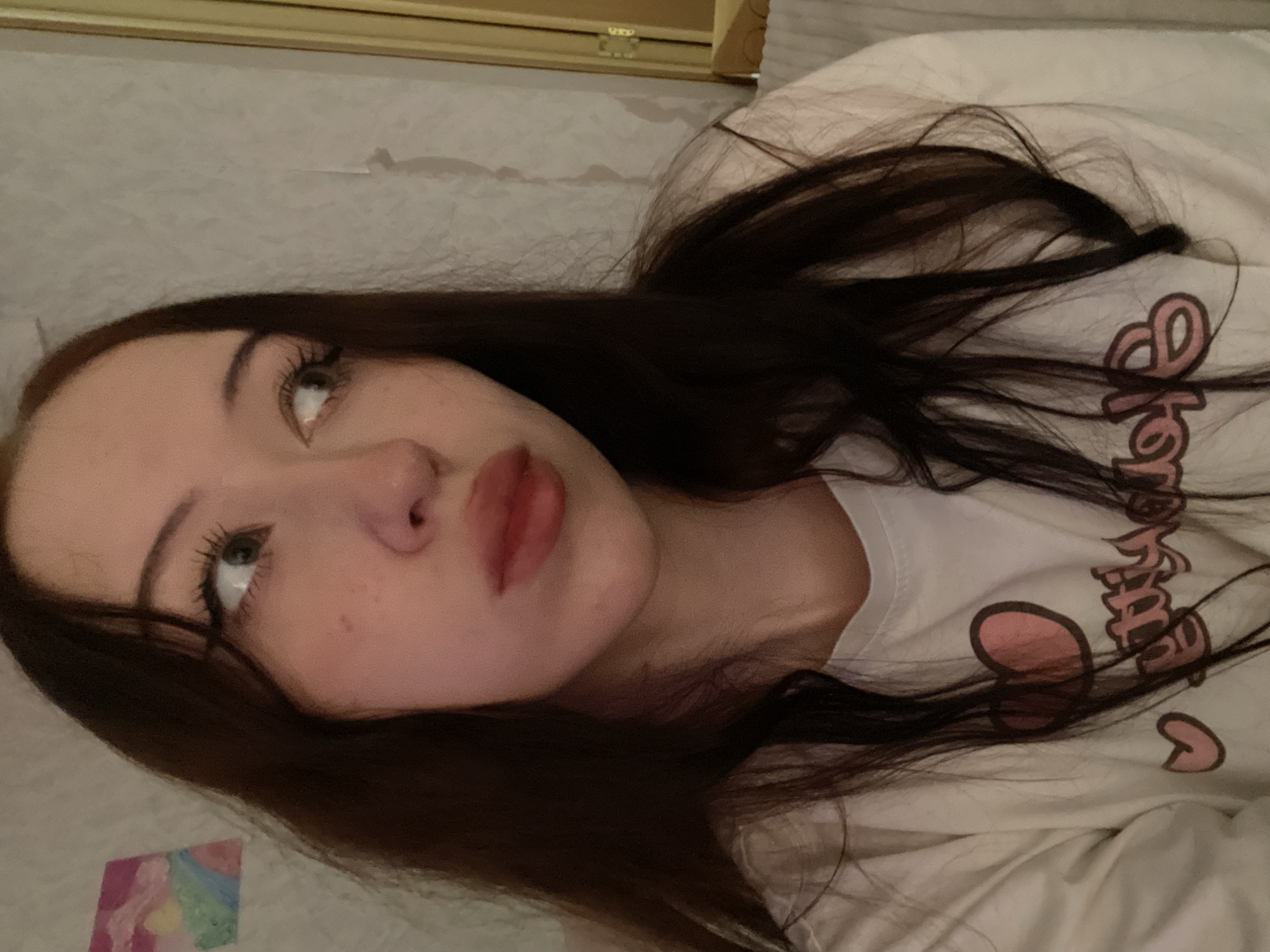 A young woman with long, dark brown hair is pictured in a close-up shot. She is wearing a white t-shirt with a pink heart and text graphic. Her gaze is directed to her right, with her mouth slightly pursed. Her skin appears fair, with some subtle blush on her cheeks. In the background, a textured, light-colored wall is visible, with a framed piece of art on the left side. The overall lighting suggests an indoor setting, possibly a room. The image does not contain any discernible elements that would indicate its location in Votkinsk, Russia, or any specific urban environment.