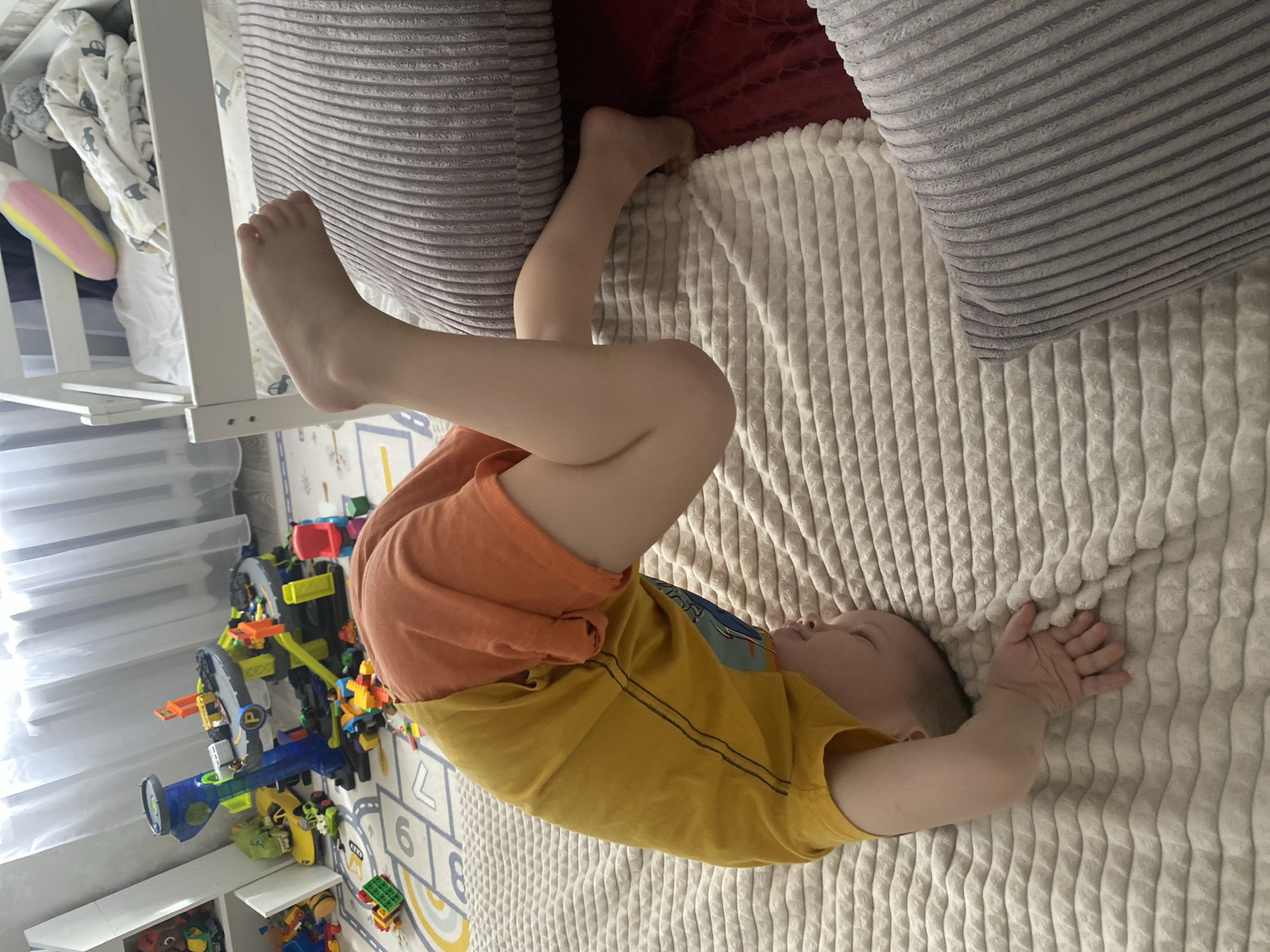 A young child is lying on a bed, angled diagonally in the frame. The child is wearing a yellow t-shirt and orange shorts. Their legs are bent, with one foot pointing upwards towards a white crib or bed frame. The bed has a textured beige blanket, and there are grey pillows at the top of the frame. To the left, there is a colorful collection of toys on the floor, including a toy track and building blocks. The floor has a play mat with numbers and letters. The location appears to be indoors, in a child's bedroom. There is no indication that this image was taken in Sochi, Russia.