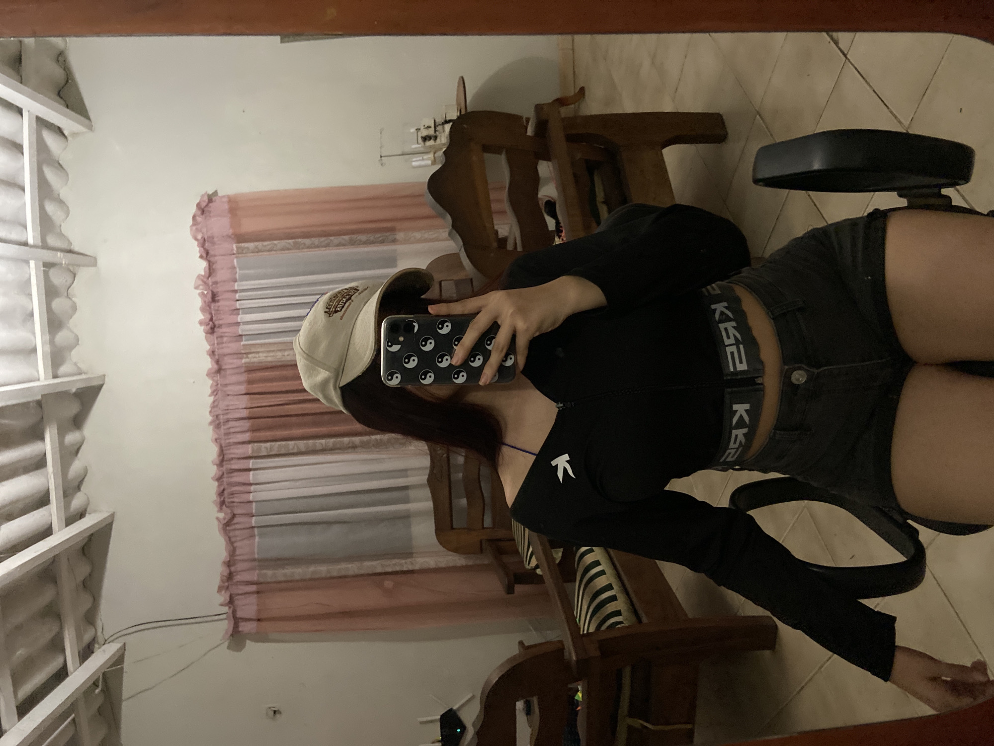 The image shows a person taking a selfie in a mirror. The person is wearing a beige baseball cap, a black long-sleeved top with a small white logo on the left shoulder, dark denim shorts, and a black belt with white text. The phone in the person's hand has a black case with a pattern of white yin-yang symbols. The background includes a window with pink and white curtains, a wooden chair, and a swivel chair. The location, Juan Griego, Venezuela, is not identifiable from the image.