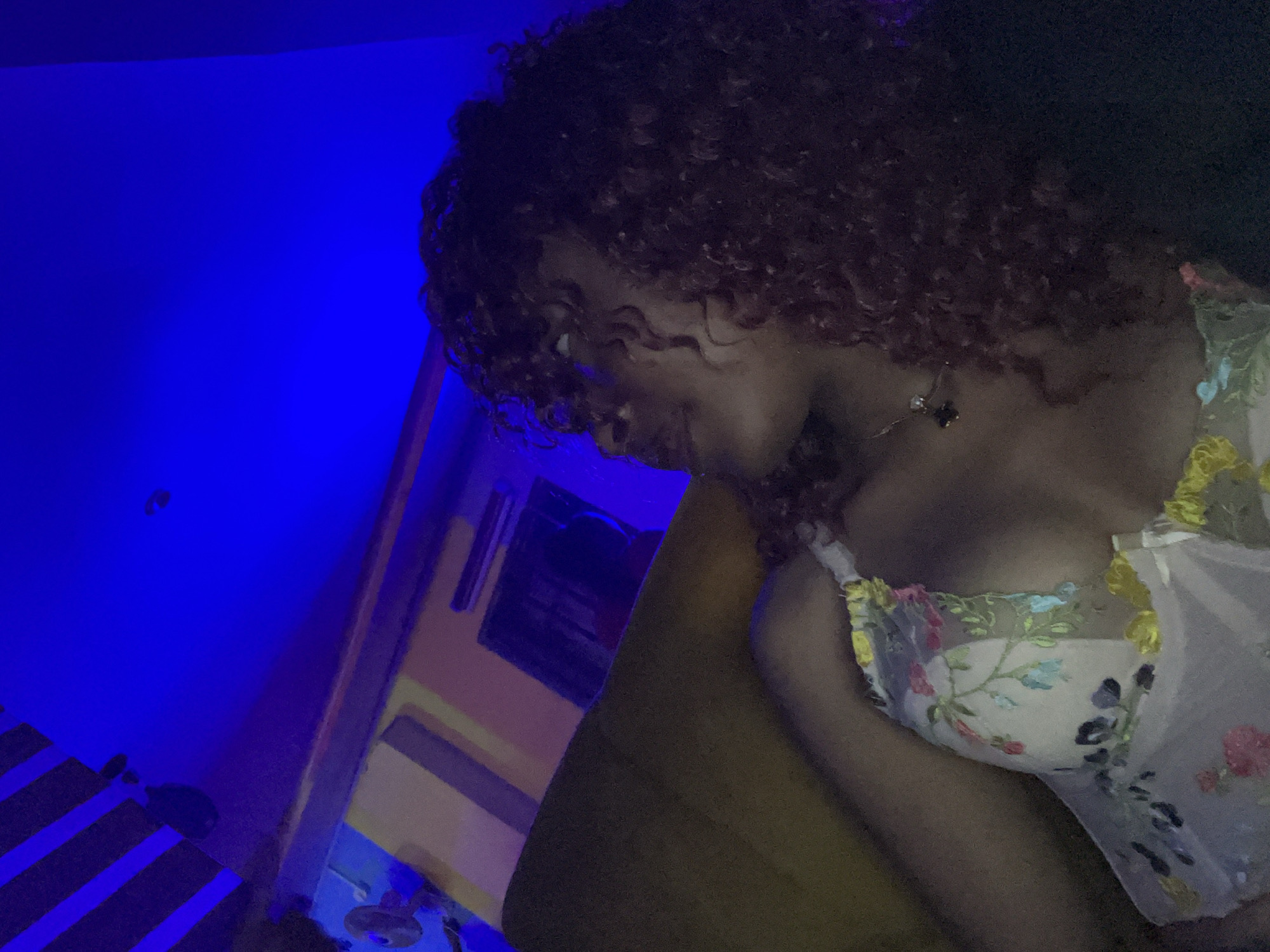A person with dark skin and curly dark hair is lying on a yellow couch, illuminated by blue light. The person is wearing a white bra with colorful floral embroidery. In the background, there are walls with striped patterns and what appears to be a shelf or ledge. The overall setting suggests an indoor environment, possibly a room with mood lighting. The location is identified as Unknown, Nigeria.