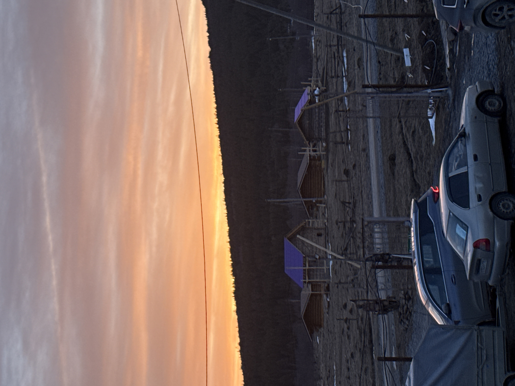 An outdoor scene in Tyulyuk, Russia, depicts a rural landscape during either sunrise or sunset. The upper sky displays a gradient from pale grey-blue at the top to vibrant orange and yellow near the horizon, streaked with thin clouds. A single dark power line spans horizontally across the upper mid-section of the sky. Below, a dense, dark coniferous forest line silhouettes a hillside against the illuminated sky.
The midground features an expanse of dry, dark earth with sparse vegetation and scattered patches of white snow. Three log cabins are visible; two possess distinctive purple tarp roofs, indicating ongoing construction or renovation. The central cabin also shows exposed wooden framework. Various wooden fences and utility poles with associated wiring are present, segmenting the terrain. A decorative metal gate is visible in the background of the vehicle area.
The immediate foreground consists of a dirt or gravel surface. A grey utility trailer, covered by a tarp, is partially visible on the left. Centrally, a dark grey sedan is parked facing right, displaying a Russian license plate (E100CK 193). Partially visible on the right, a dark grey SUV is parked behind the sedan, also facing right, with its left rear tail light illuminated red. No individuals are present in the image.