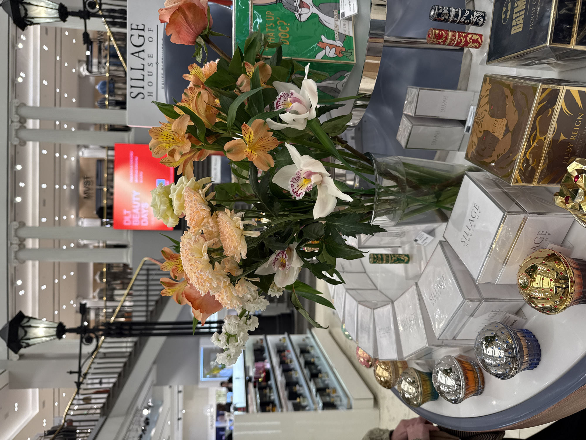 The image shows a display of luxury perfumes and cosmetic products on a white, curved counter. In the foreground, a vase filled with a vibrant arrangement of peach-colored chrysanthemums, orange alstroemeria, and white orchids is prominently placed. Several decorative, egg-shaped perfume bottles with reflective gold and silver surfaces are arranged on the counter. Behind the flowers, white boxes with "SILLAGE HOUSE OF" branding are stacked. To the right, a green box featuring a Bugs Bunny illustration with the text "WHAT'S UP DOC?" is visible, along with smaller cylindrical perfume bottles. In the background, a multi-story interior of a department store is depicted, with escalators, lamps, white columns, and shelves stocked with various products. A bright red digital sign with "DLT BEAUTY DAYS" text is also visible. The overall scene suggests a high-end retail environment. The context of Saint Petersburg, Russia, is not visually evident within the image itself.