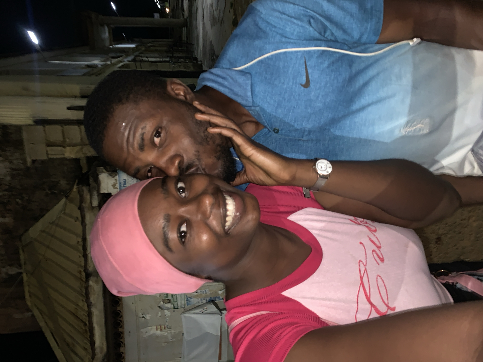 Two dark-skinned individuals, one male and one female, are posing closely together for a photo outdoors. The woman is in the foreground, wearing a pink headscarf and a pink and white shirt. She is smiling. The man is behind her, wearing a blue Nike shirt and a wristwatch. He appears to be kissing her cheek. The location seems to be a narrow street or alley in Jos, Nigeria, based on the architectural style and environmental details in the background. There are building structures and an outdoor setting, and the illumination suggests it's nighttime.