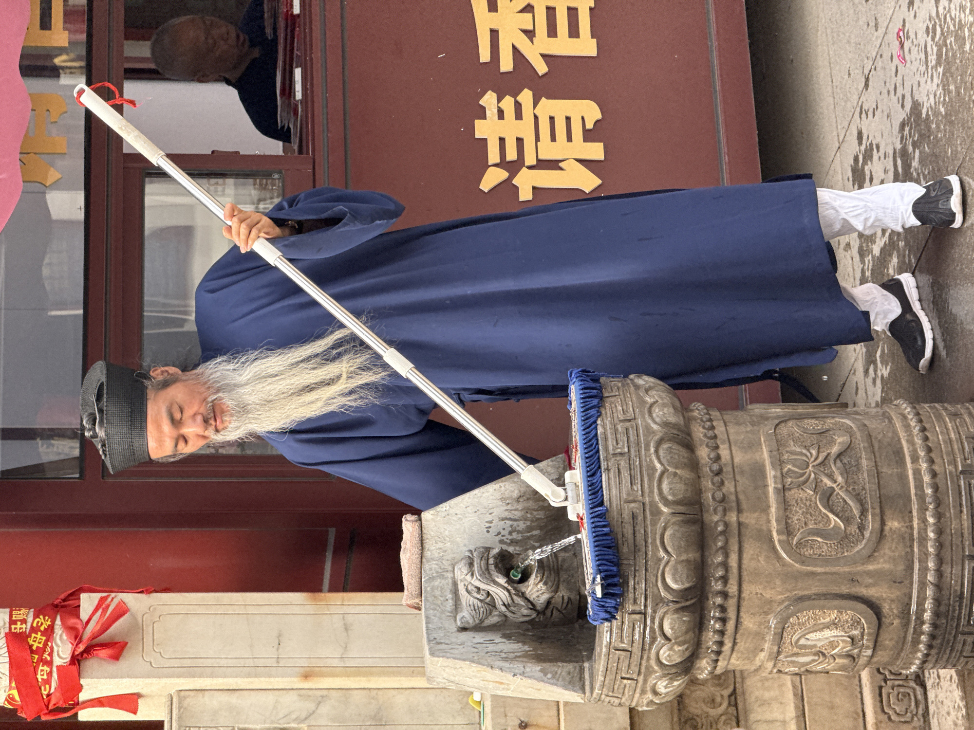 A person in a dark blue robe and black cap is using a blue and white mop on a stone pillar in Lintong District, China. The person has a long white beard and is wearing white pants and black-and-white sneakers. The stone pillar is decorated with carvings and is dispensing water. Behind the person are red buildings with gold lettering and another person looking out of a window. The ground is wet.