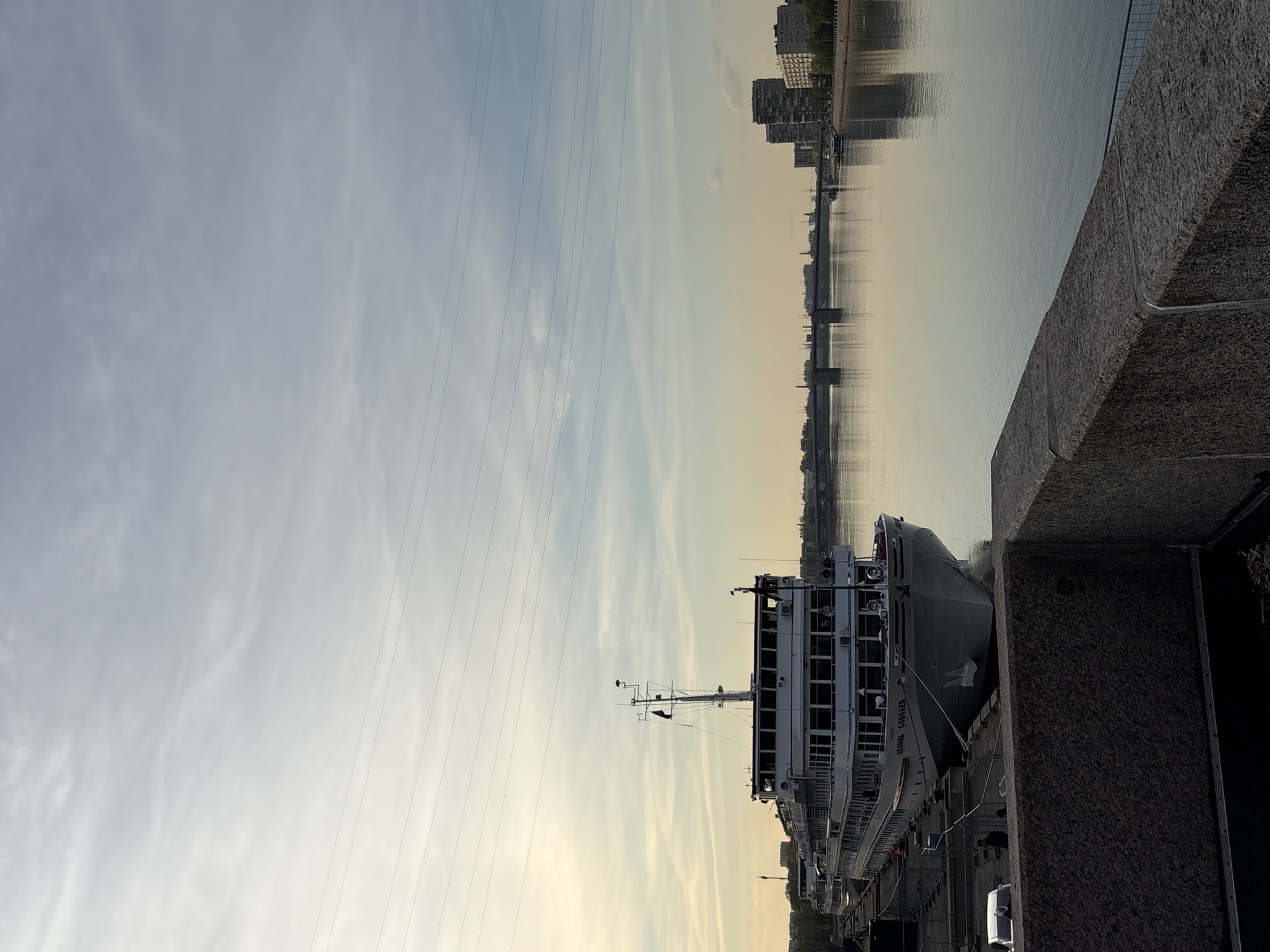 The image depicts a docked passenger ship, likely on the Neva River in Saint Petersburg, Russia. The ship is multi-decked with visible windows and a mast. A stone embankment is visible in the foreground. The river reflects buildings on the opposite bank, including a bridge. Power lines stretch across the sky, which is a light blue with some cloud cover. The light suggests either sunrise or sunset. The ship has the name "LENINA COBOLEV" visible on its side. The image appears to be taken from a standing position on the embankment.
