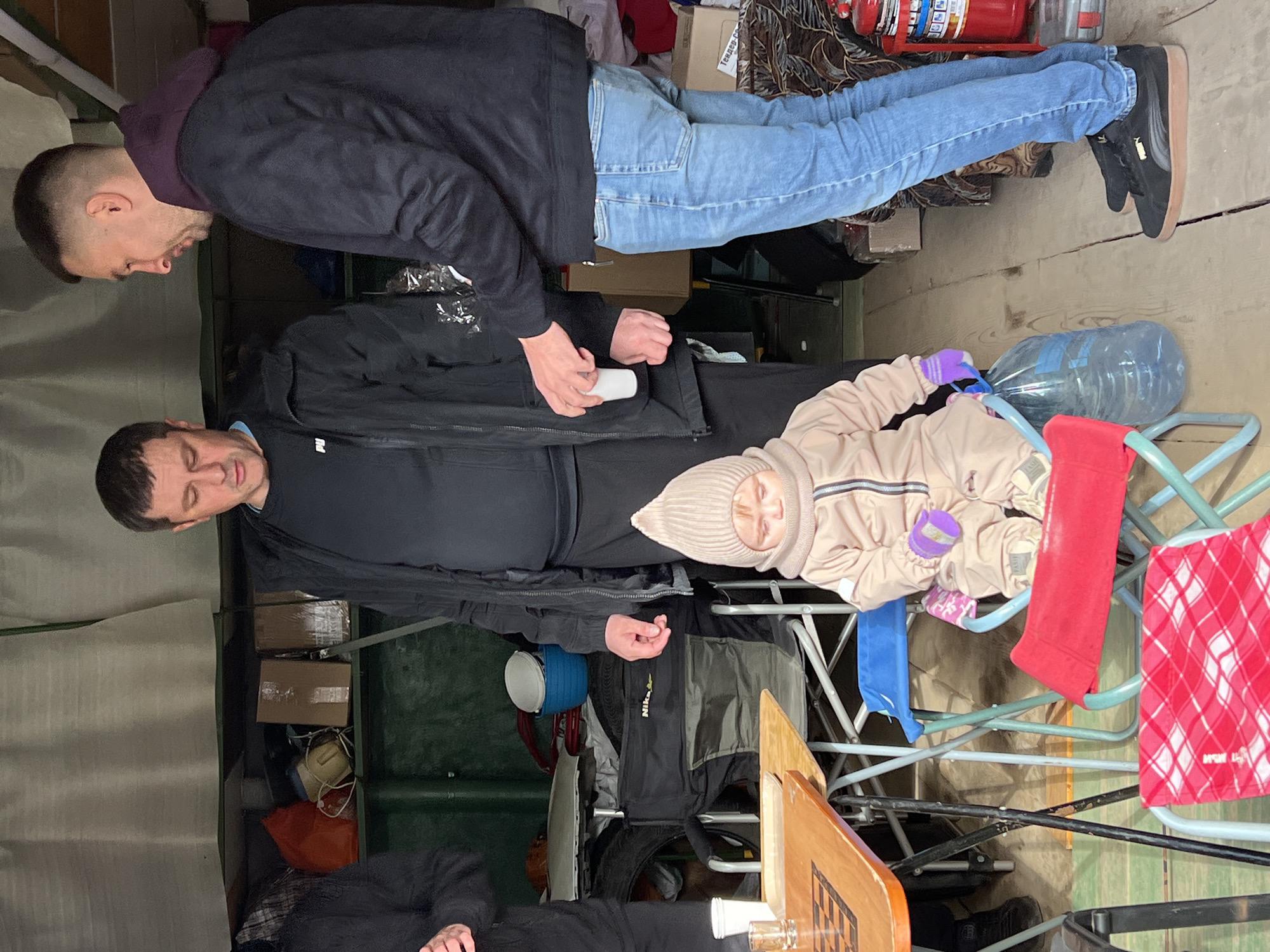 The image depicts three individuals in what appears to be a cluttered interior space, potentially a garage or storage area. An adult male with a short haircut is standing, wearing a dark hooded sweatshirt, light blue jeans, and black sneakers. He is partially cropped in the image. A second adult male, wearing a dark jacket over a dark shirt, stands closer to the center, holding a small white object in his hands. Seated on a blue and red folding chair is a child, dressed in a beige snowsuit and hat, with purple mittens. A large clear plastic bottle of water is positioned near the child. The surrounding environment is filled with various objects, including boxes, bags, a fire extinguisher, and other miscellaneous items. The location is specified as Noviy, Russia.