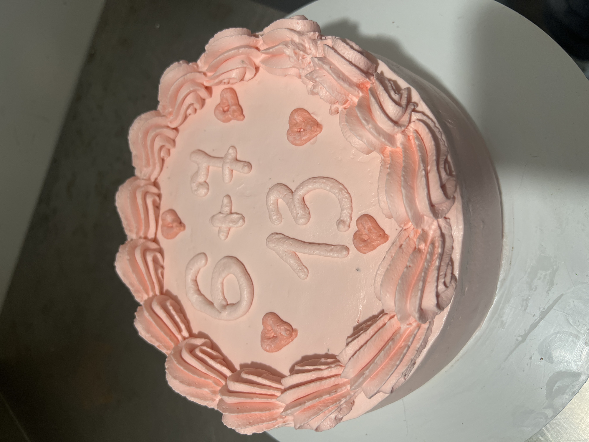 A top-down view captures a small, round cake decorated in varying shades of pink. The cake's surface is a smooth, light pink frosting, adorned with the numbers "6" and "13" separated by a plus sign "+", all rendered in the same pink frosting. Small, stylized heart shapes, also in pink frosting, are scattered around the numbers. The edge of the cake is decorated with a textured, darker pink frosting pattern. The cake sits on a white, circular cake stand or surface. The background is a combination of white and a darker gray surface.
The location in the city of Chaikovsky, Russia is not discernible from the image.