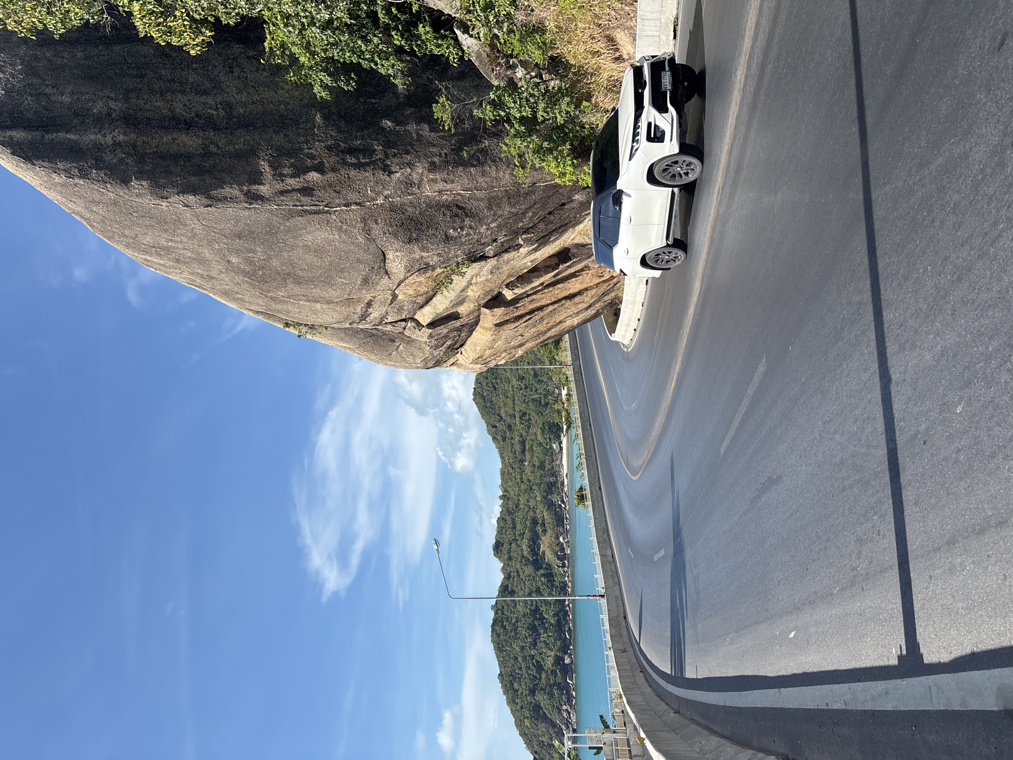 The scene depicts a road in Baan Chaweng Noi, Thailand, flanked by a large rock formation on one side and the ocean with a forested hillside on the other. A white SUV is parked on the roadside next to the rock. A lamppost is visible along the road, casting a shadow. The sky is blue with some clouds. The road curves around the rock formation.