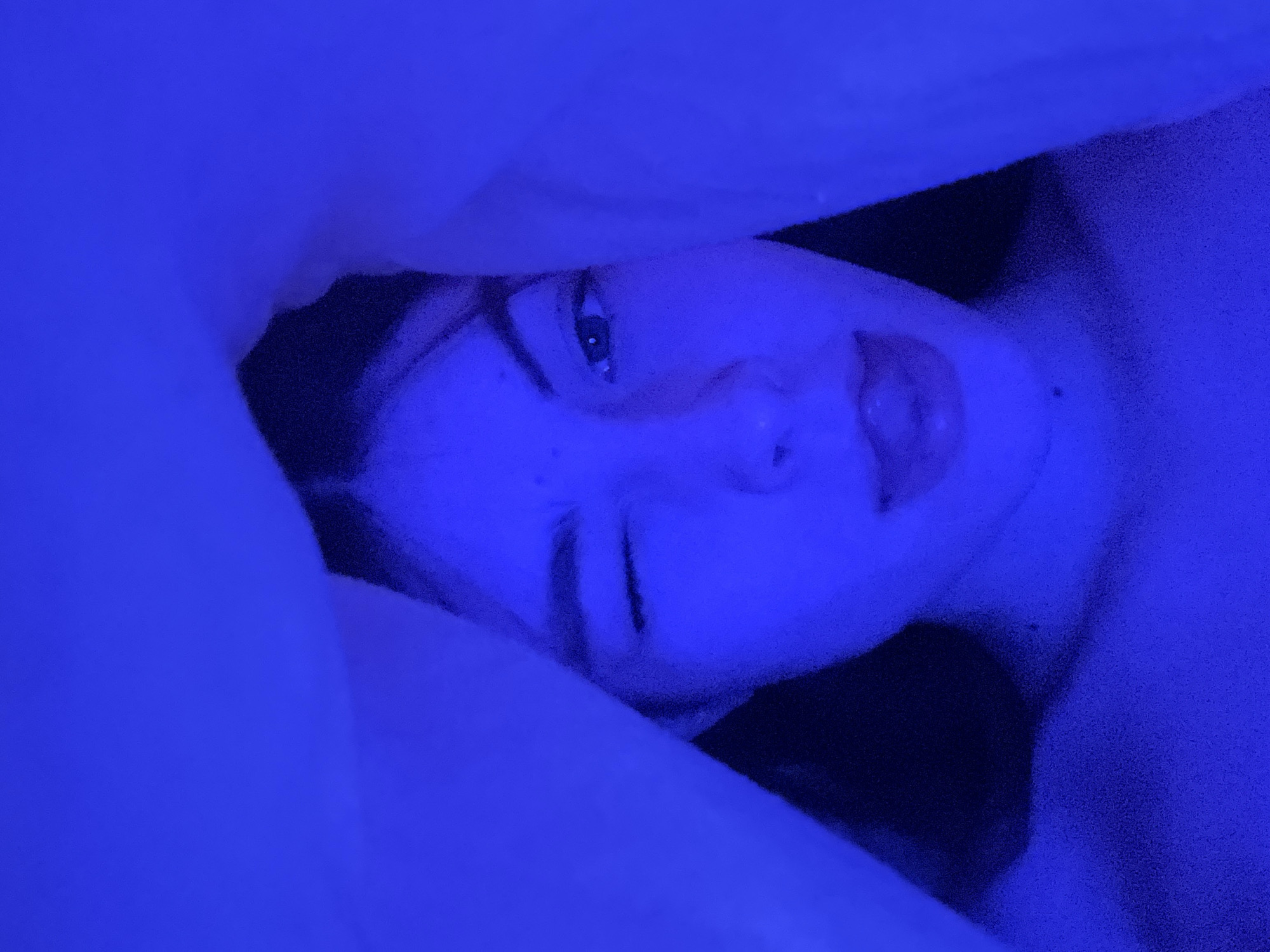 The image presents a close-up, angled portrait of a person, likely a young female, lying down in a deeply blue-lit setting. The subject's face is positioned centrally and slightly to the right, framed by what appears to be a white pillow or blanket that occupies the left and upper portions of the frame.
The person's right eye (from the viewer's perspective) is open and looking directly towards the camera, while the left eye is partially or fully closed, suggesting a wink or a blink. Their lips are slightly parted, revealing a hint of the tongue. The face shows faint details like freckles, illuminated by the strong blue light. The area behind the subject's head is shrouded in deep shadow, making it indistinct.
The entire scene is saturated with a striking blue hue, indicating either very dim ambient lighting with a blue source (like an LED light strip or a lamp with a blue filter) or a heavily color-graded photograph. This monochromatic blue lighting creates an intimate, dreamy, or nocturnal atmosphere, suggesting the scene takes place indoors, possibly in a bedroom during the evening or night. The expression conveyed is one of relaxation, playfulness, or gentle engagement.
No specific objects or details within the image directly indicate a geographical location, though the provided context suggests Saint Petersburg, Russia. There is no visible text in the image.