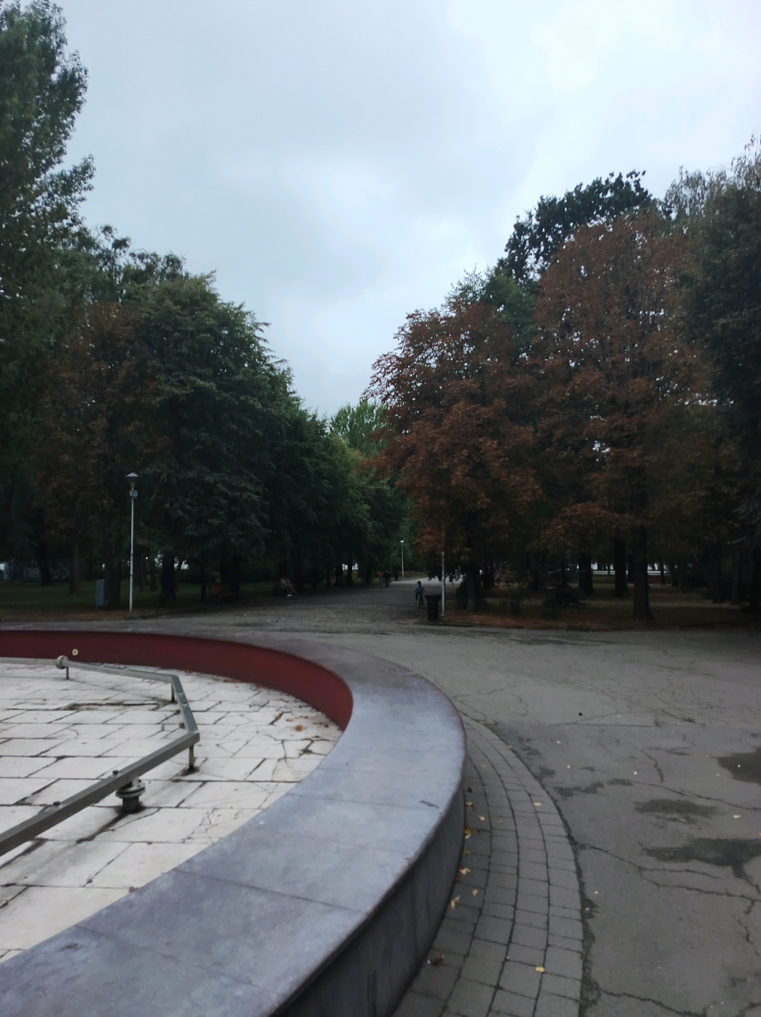 The image depicts a park in Ternopil, Ukraine, under an overcast sky. In the foreground, a grey, curved structure, possibly the edge of a fountain or plaza, is visible. To the left, a tiled area with a metal railing is also present. Beyond this, a paved pathway leads into the park, lined with trees exhibiting autumn foliage in shades of green and brown. A lamppost stands near the pathway. Further into the park, a line of trees forms a dense backdrop, with some people seated on benches in the distance. The overall atmosphere is subdued, consistent with a cloudy day in autumn.