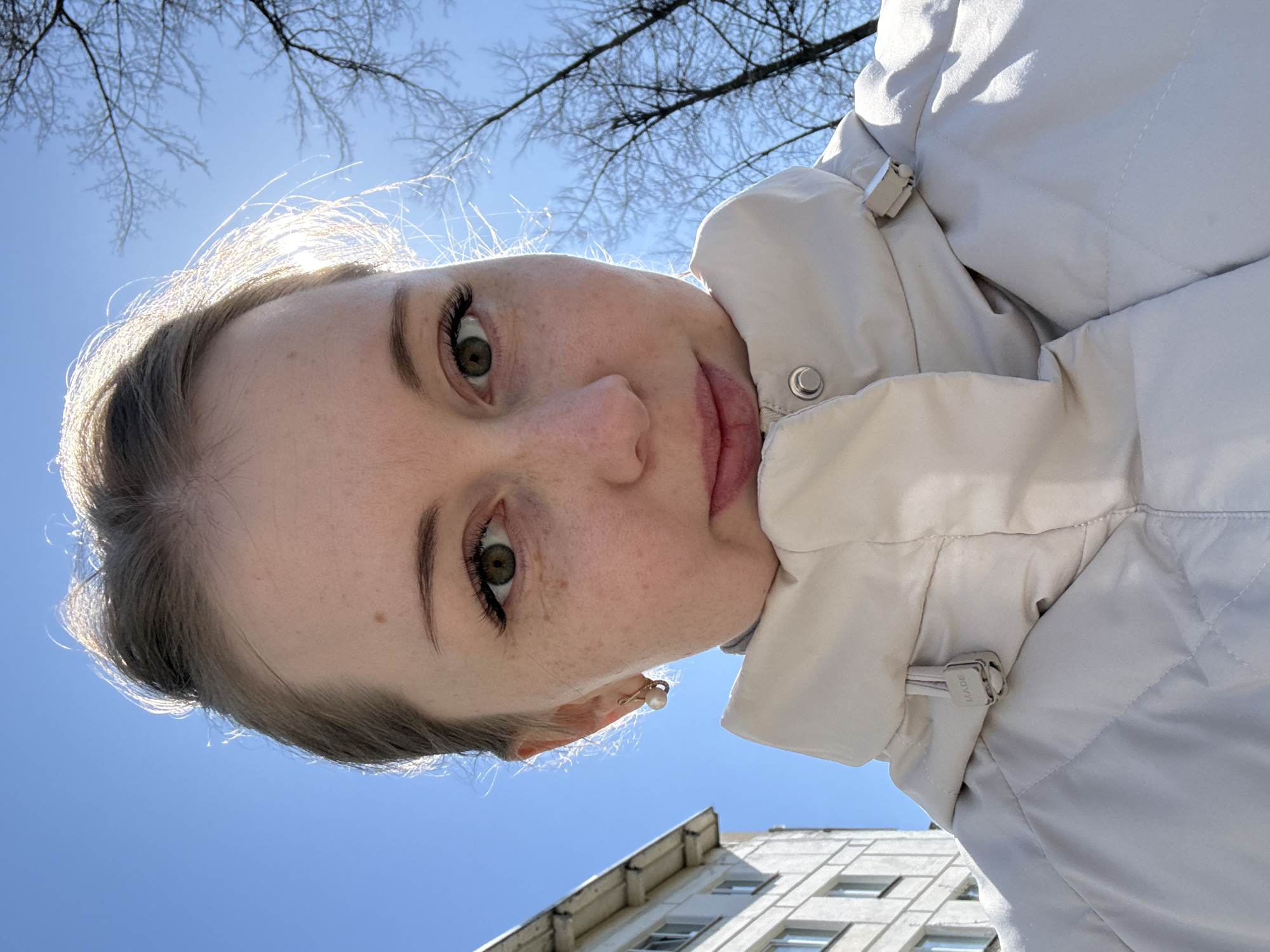 A photograph taken from a low angle depicts a person with fair skin and light brown hair, framed against a clear blue sky and the upper branches of bare trees. The person's face is prominent, displaying green eyes and freckles. They are wearing a light-colored padded jacket with a high collar. In the background, a building with multiple windows is visible. The location is an urban environment, though the specific city or region is unknown.