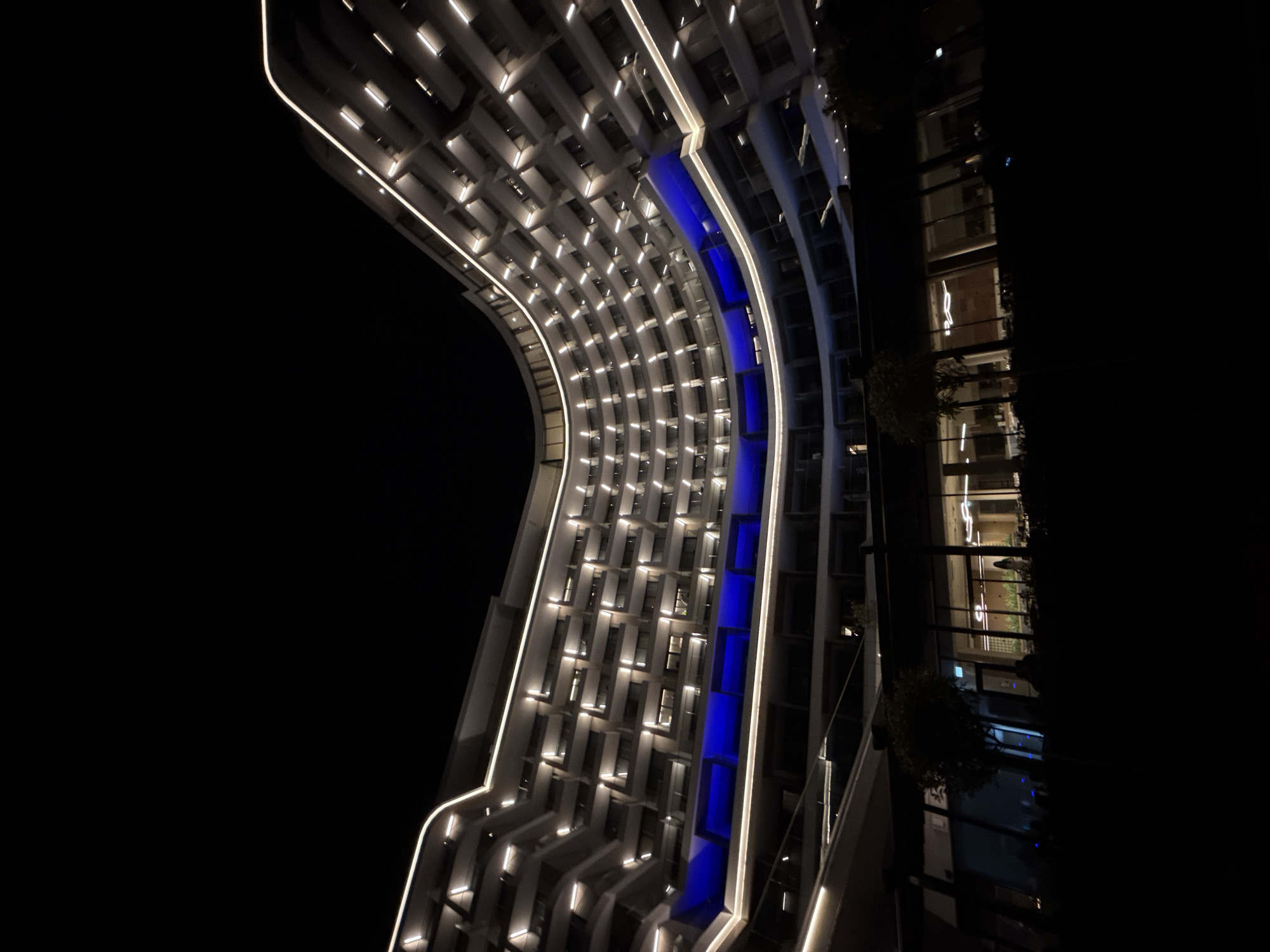 This media file captures a modern architectural building at night in Varna, Bulgaria. The main subject is a striking curved building with a facade illuminated by numerous lights, creating a sense of depth and complexity. White linear lights trace the contours of the building, accentuating its unique architectural design. A prominent vertical strip of vibrant blue light runs down the central section of the curved facade, adding a dramatic and futuristic element.
The building appears to be residential or commercial, with many rectangular windows visible, each lit from within, suggesting occupancy. To the right, a section of the building is clad in dark glass, reflecting the surrounding darkness and the interior lights, giving glimpses of internal spaces and foliage. The time of day is clearly night, with the stark blackness of the sky contrasting sharply with the illuminated architecture. The weather appears clear, as there are no visible atmospheric obstructions.
The overall mood conveyed by the image is one of modern elegance and sophisticated urban design. The lighting creates a visually captivating scene, highlighting the building's form and its integration with the night environment. There are no people or specific activities depicted, focusing solely on the architectural subject. No visible text is present in the image. The curved design and the interplay of light suggest this might be a landmark or a notable structure within Varna.