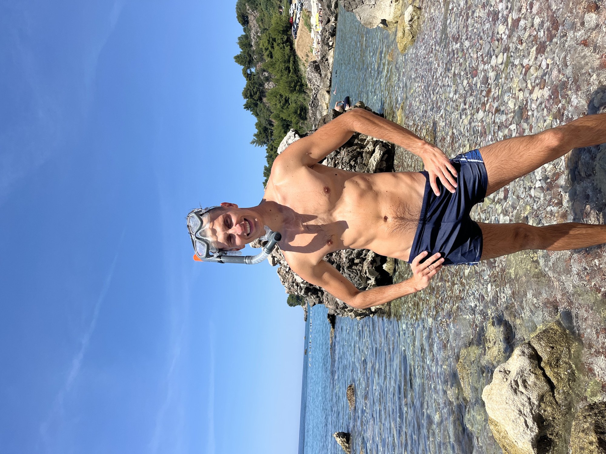A young Caucasian man stands waist-deep in the clear, shallow water of a rocky beach in Sutomore, Montenegro. He is wearing dark blue swim trunks and a snorkeling mask with a snorkel attached to the top of his head. The man is smiling and looking directly at the camera with his hands on his hips. The water is transparent, revealing the colorful pebbles and rocks on the seabed. In the background, a large rock formation is visible, with green trees and vegetation covering a hillside beyond. The sky is a clear, bright blue. Other beachgoers and parked cars can be seen in the distance.