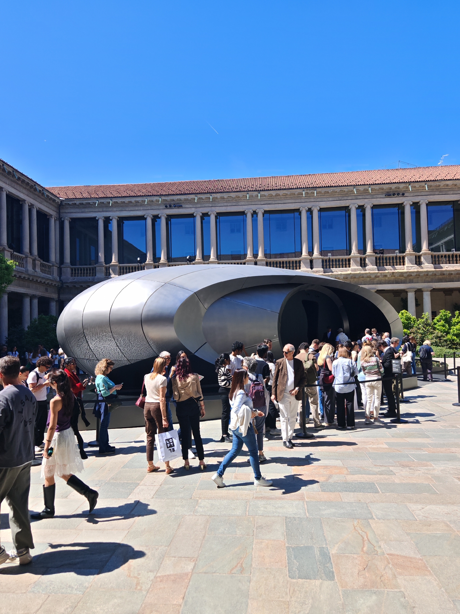 The image shows a modern, silver, pod-like structure situated in the courtyard of a classical building, likely a museum or public building. The building is two stories high, constructed with columns and large windows that reflect the sky. A crowd of people is gathered near the entrance of the silver structure, suggesting it's an exhibit or attraction. The ground is paved with stone slabs. The setting appears to be a city, possibly Milan, Italy, based on visual cues of architecture and style. The sky is clear and blue, indicating a sunny day.