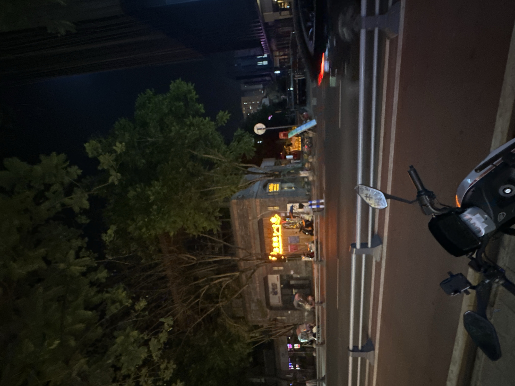 This is a nighttime, eye-level, medium shot of a street scene in the Chongqing High-tech Zone, China. In the foreground, the handlebars and front section of a black scooter are visible, parked on a sidewalk. The scooter is facing left. Beyond the scooter, a two-lane road is visible with white lane markings and a metal guardrail separating it from the sidewalk. Several cars are present on the road, with their headlights and taillights illuminated. On the opposite side of the road, there is a building with a brick facade and illuminated windows. Trees with sparse foliage line the street. In the background, taller buildings with illuminated windows are visible, suggesting a developed urban area. The overall atmosphere is dark, with artificial lighting from streetlights and buildings illuminating the scene.