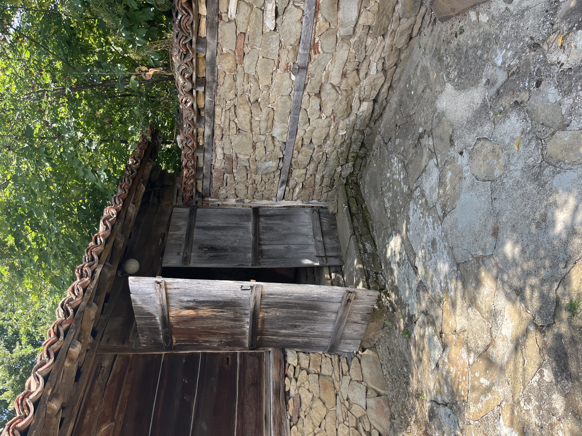 The image captures an exterior view of a traditional Bulgarian house in Zheravna, Bulgaria. The house features a wooden facade and a tiled roof. In the foreground, a stone wall encloses a courtyard paved with irregular stone slabs. The courtyard is partially shaded by trees. Two large wooden doors, one slightly ajar, are visible on the side of the house, leading into a dark interior. The overall scene depicts an old, rustic architectural style.
