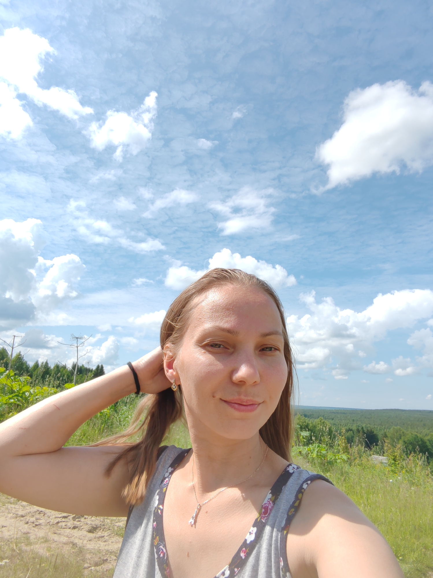 A young Caucasian woman with shoulder-length blonde hair is taking a selfie outdoors. She is wearing a gray tank top with a floral pattern and a delicate necklace with a small pendant. Her left arm is raised, and her hand is resting on the back of her head, adjusting her hair. She is smiling slightly, looking directly at the camera.
The background suggests a rural or natural setting, possibly on a hill or elevated area. Lush green vegetation and trees are visible, extending into a distant, rolling landscape under a bright blue sky filled with white, puffy clouds. The overall impression is one of a sunny day in a scenic, natural environment. There are no visible man-made structures or urban elements that would specifically place the location within the city of Чухлэм, Russia. The environment appears to be a natural landscape.